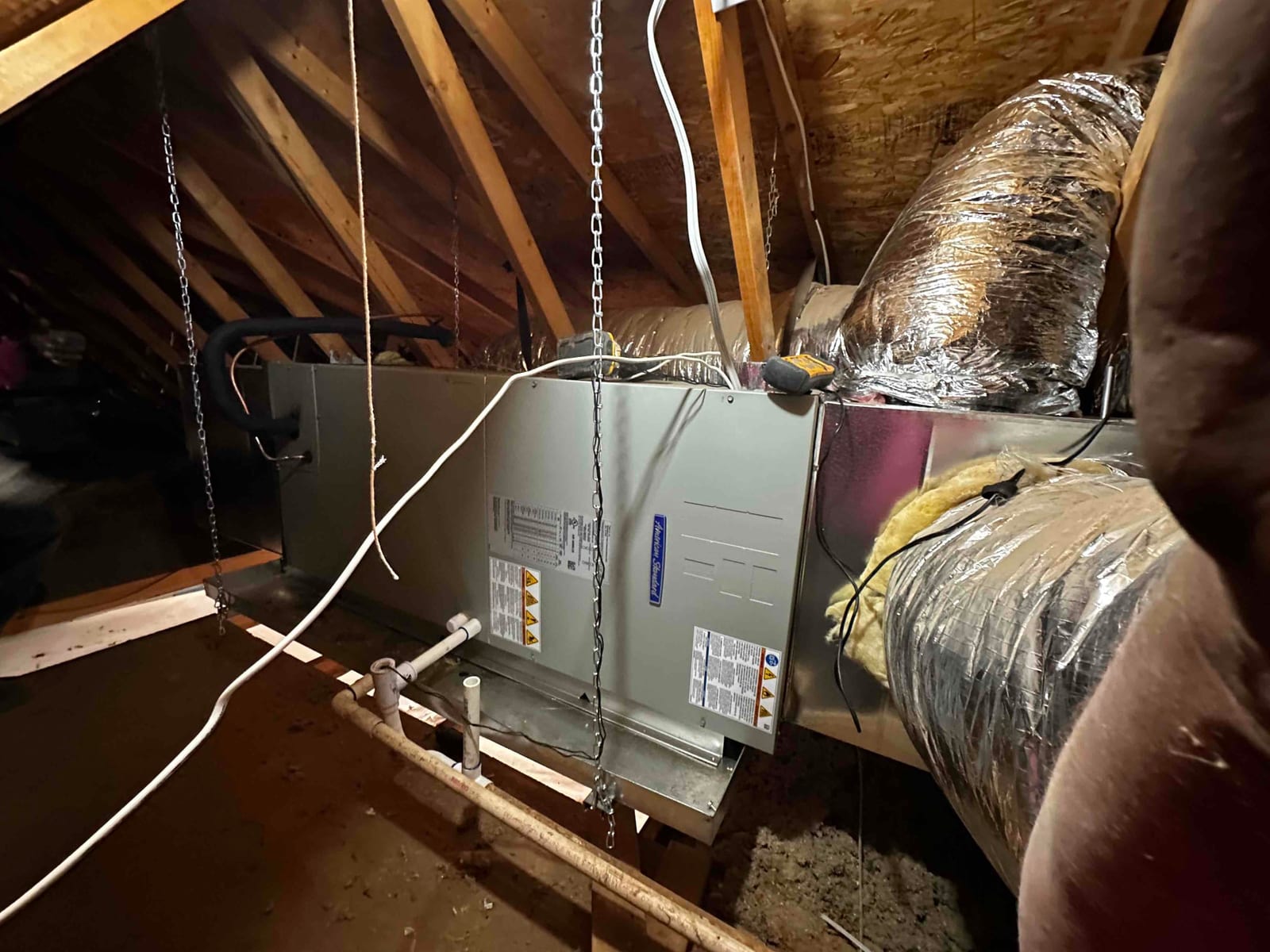 Air handler suspended in attic with connected insulated ductwork and condensate drain piping visible along the drain pan.