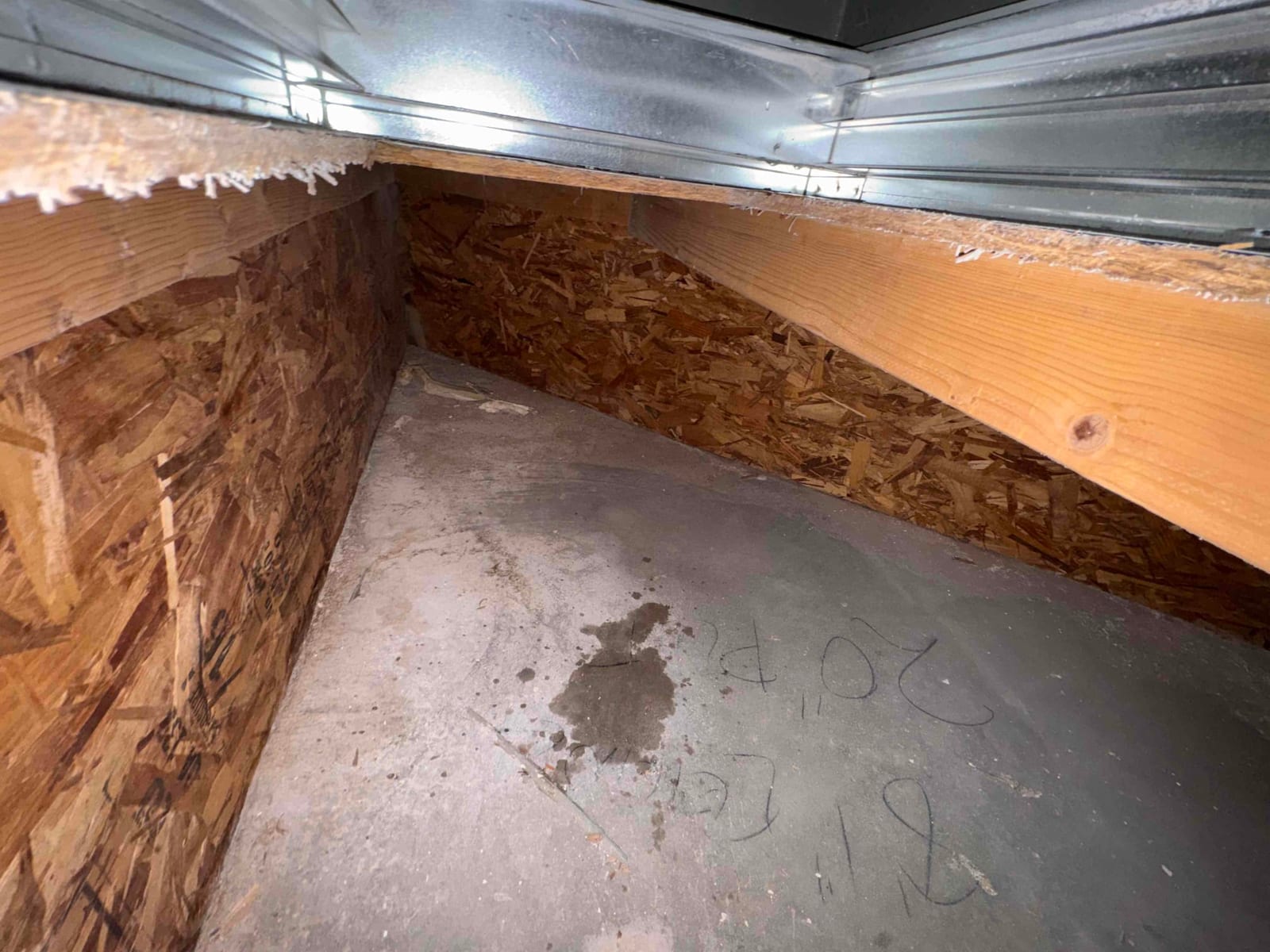 Attic or crawlspace area beneath ductwork showing a damp spot on the concrete slab near insulation and framing.