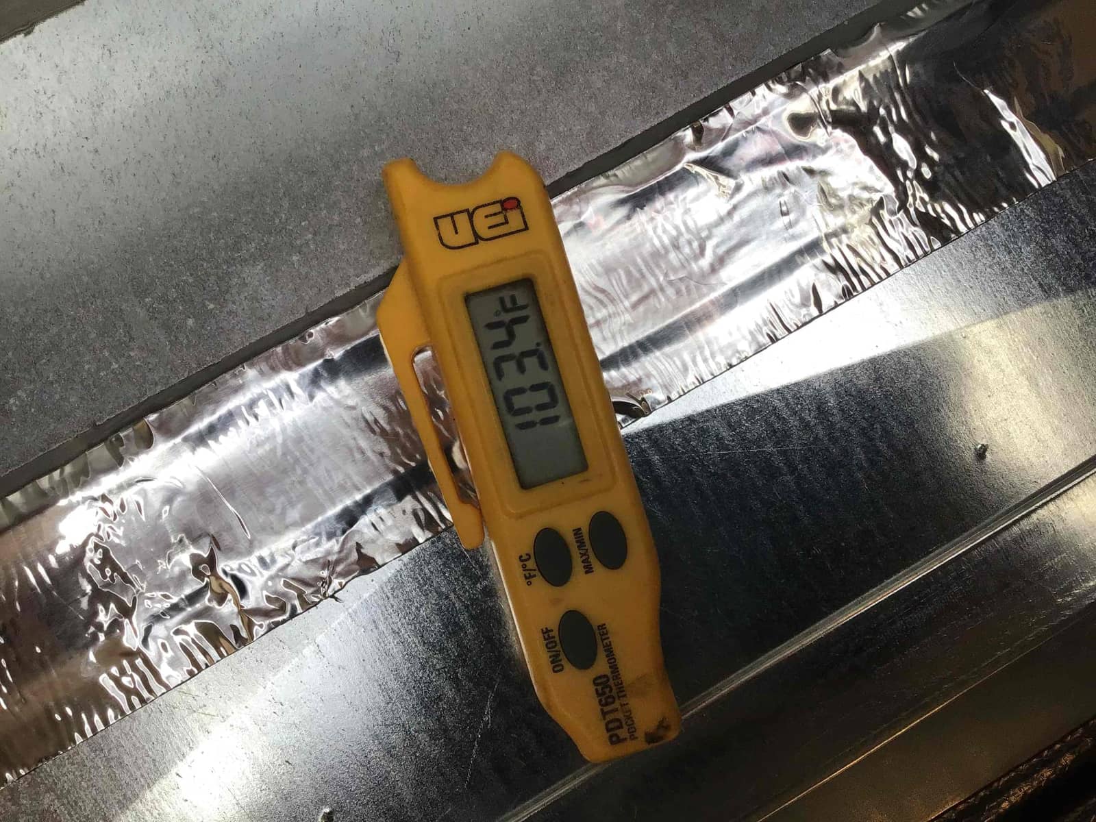 Digital probe thermometer resting on sheet metal duct shows 103.4°F, documenting air temperature during commissioning.