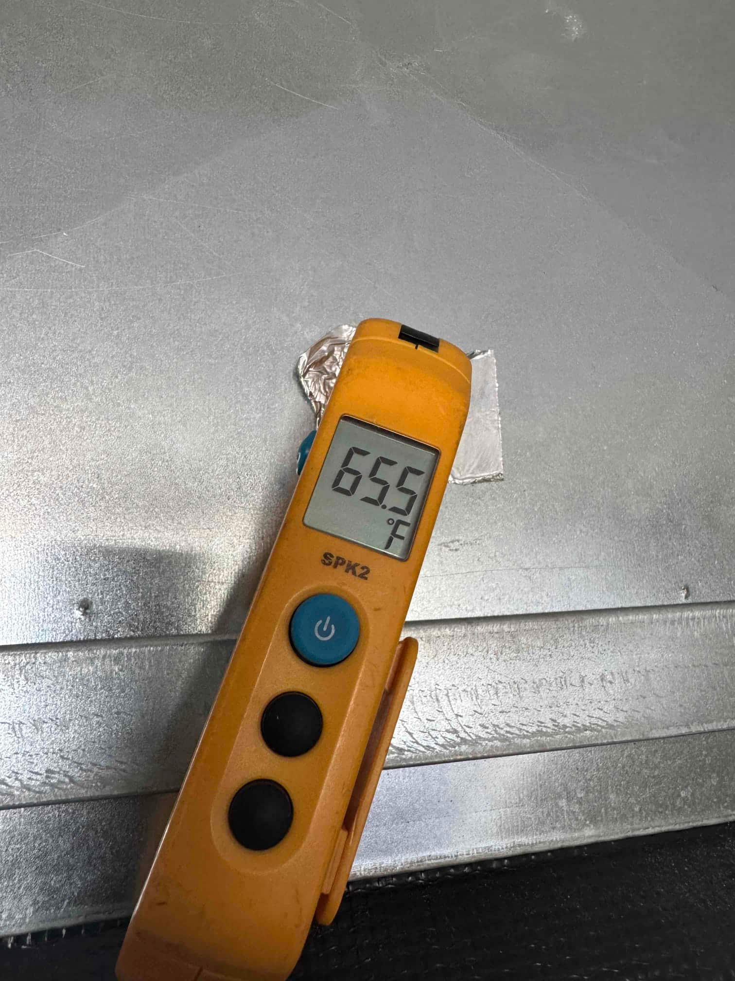 Digital thermometer shows 65.5 °F while measuring air temperature at the exterior surface of a metal duct.