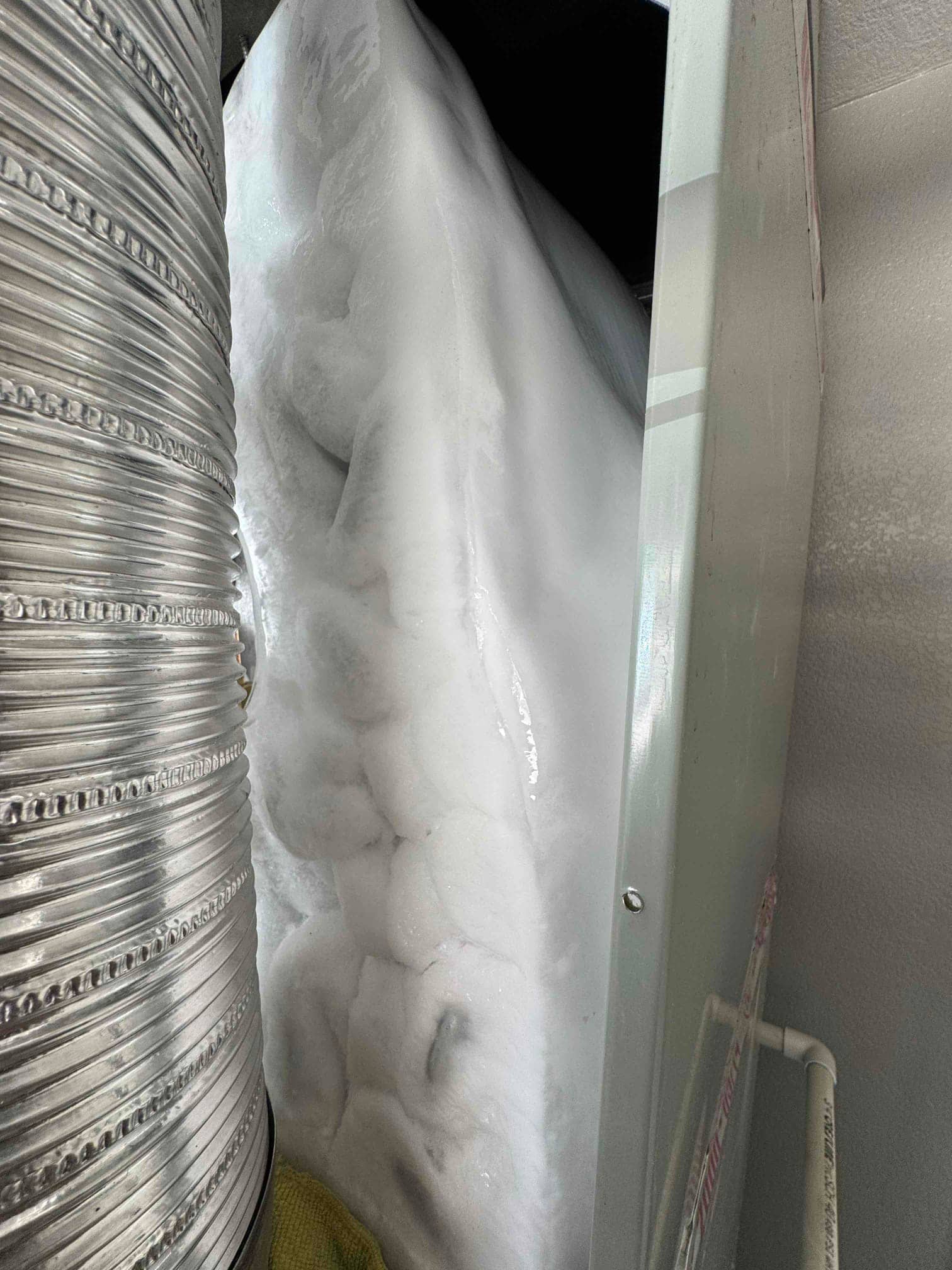 Evaporator coil housing packed with heavy ice buildup adjacent to the air handler and insulated duct connection.