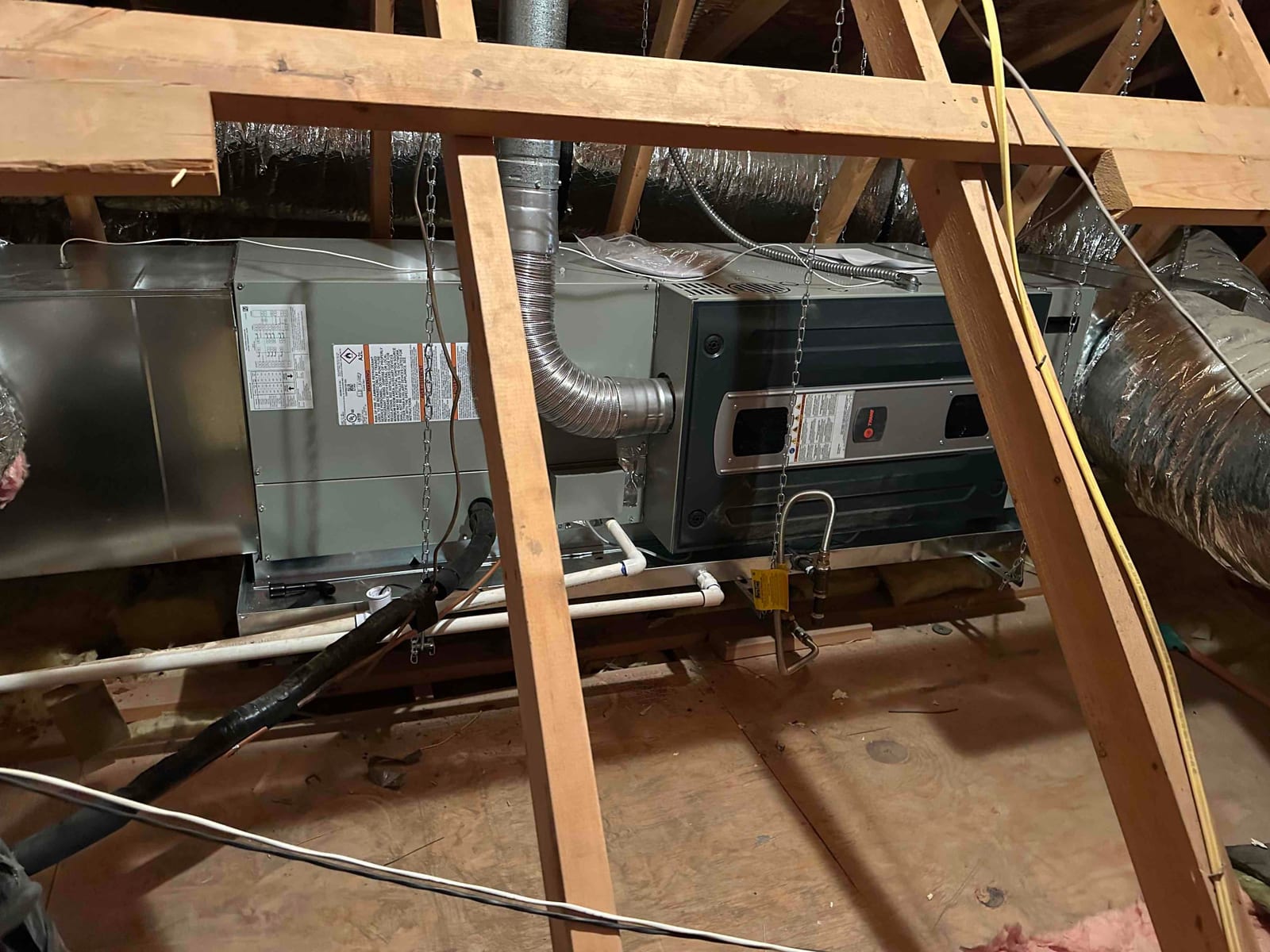 Horizontal air handler and furnace assembly suspended in attic, connected to insulated ducts, condensate drain, gas vent, and refrigerant lines.