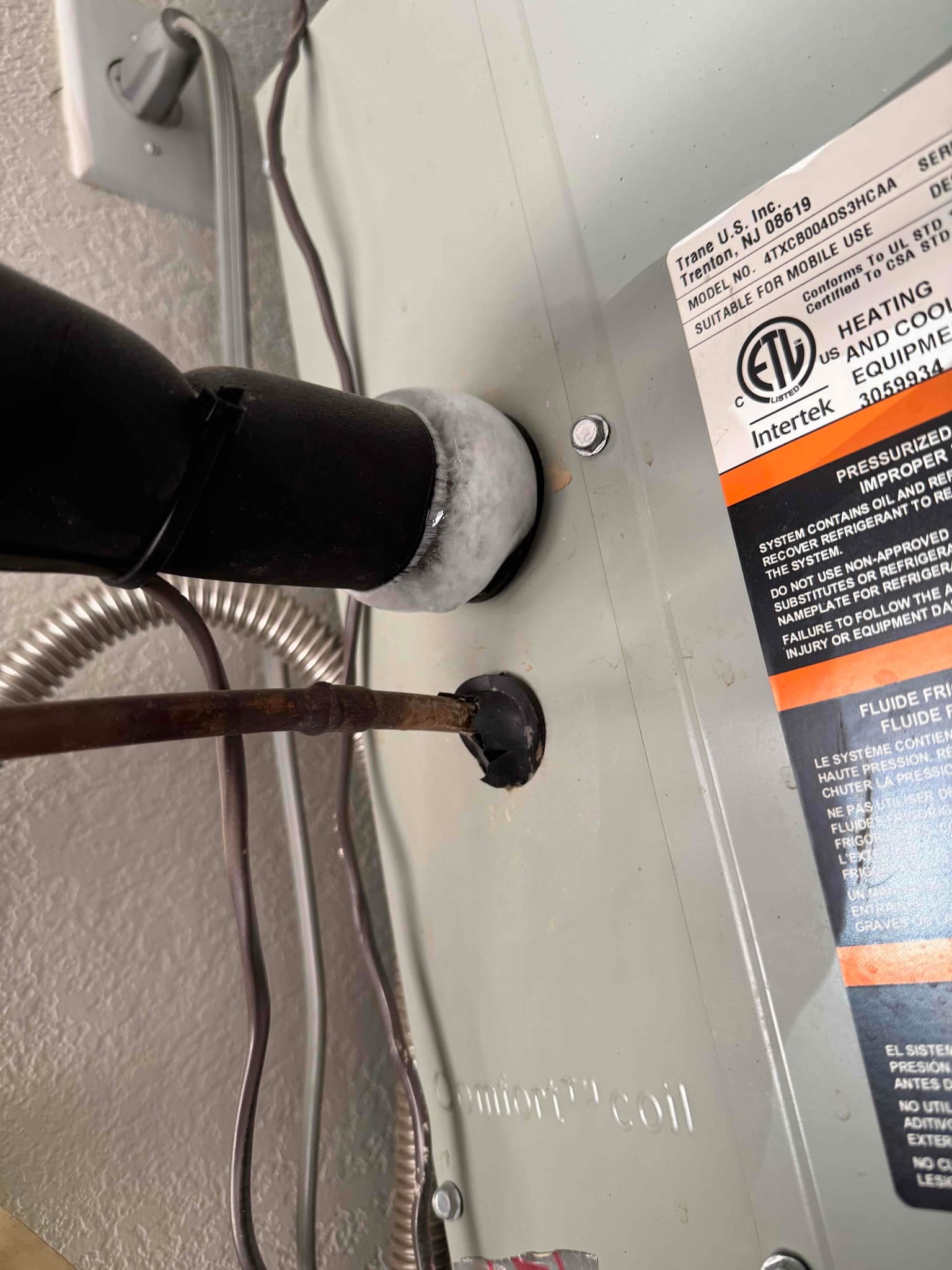 Indoor air handler cabinet with insulated drain or vent line frosted at wall penetration, showing condensation and frost on the pipe.