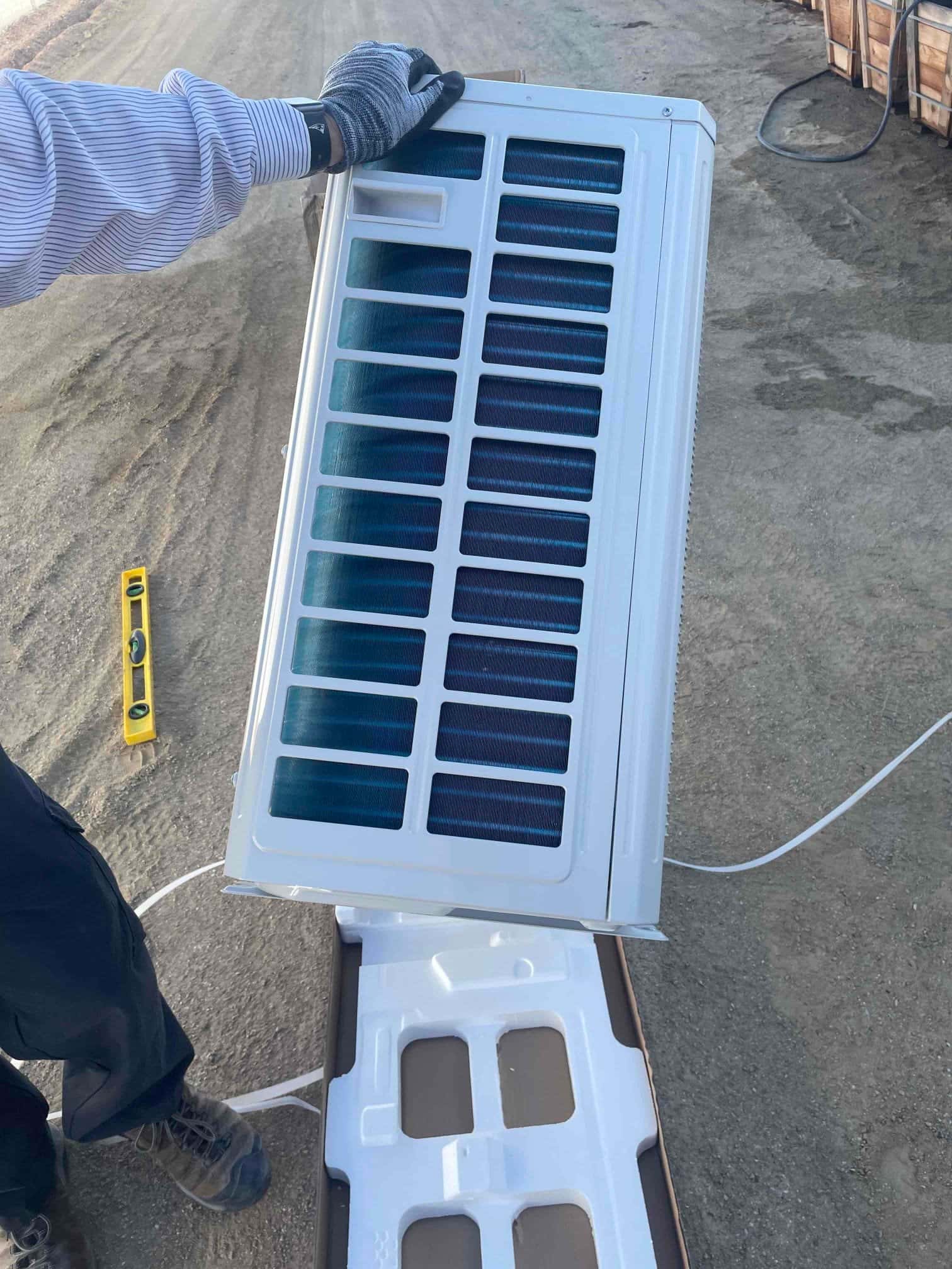 Technician unboxes and handles a new mini-split outdoor unit coil assembly at the installation site, with packaging materials on the ground.