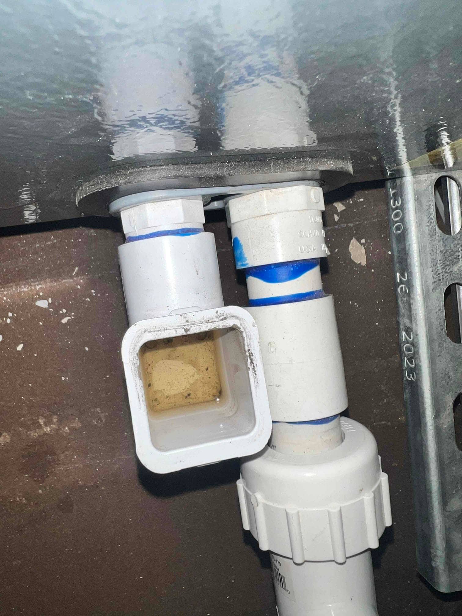 Condensate drain assembly under an indoor unit, showing PVC fittings, cleanout opening, and trapped water in the cleanout.