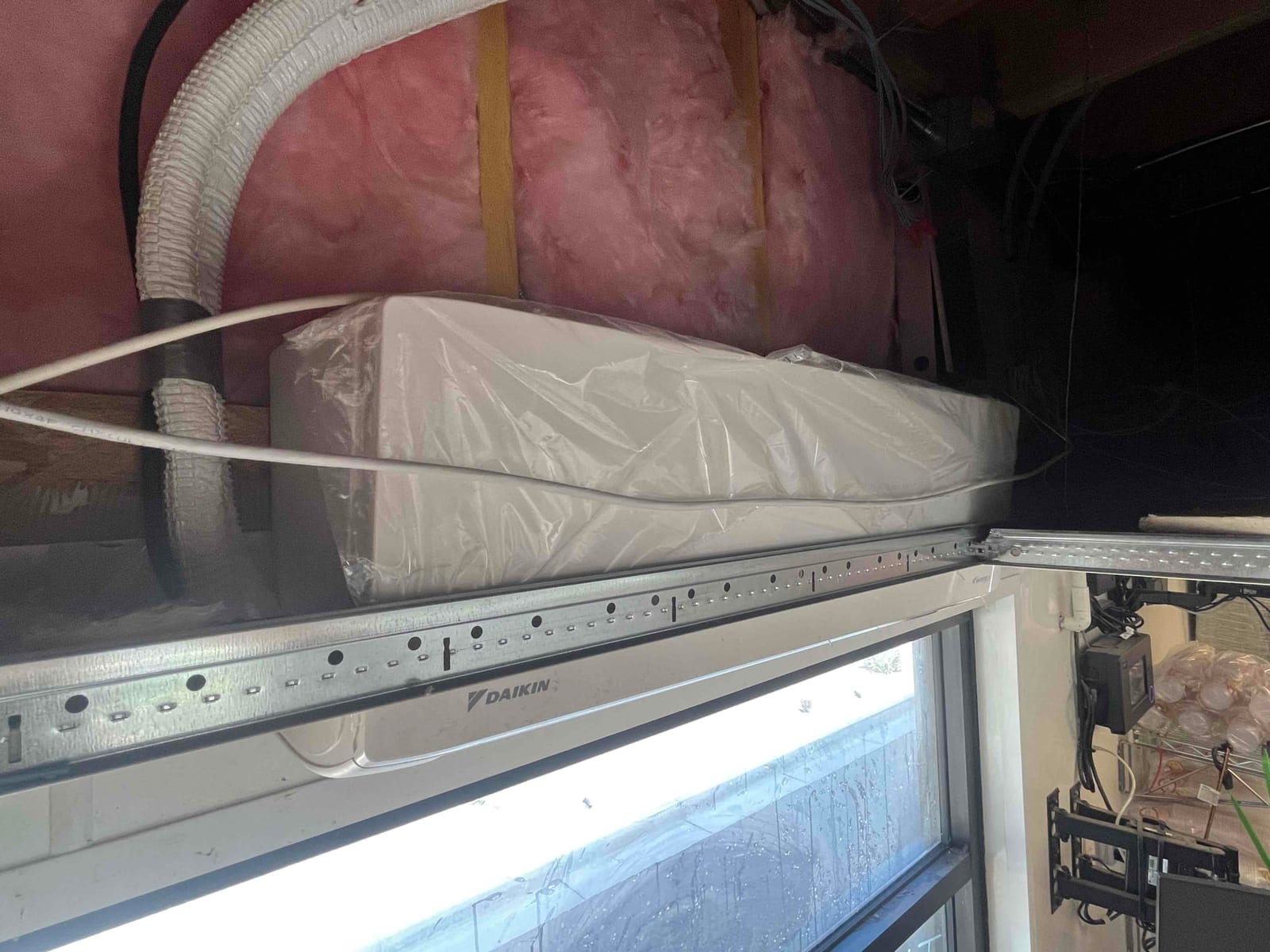 Indoor ductless air handler installed above a window, wrapped in protective plastic, with refrigerant lines and electrical conduit routed overhead.