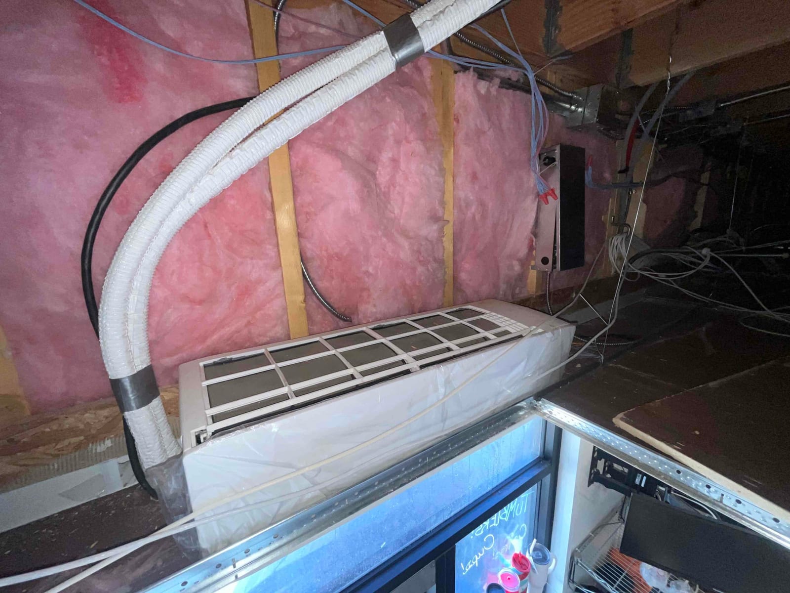 Indoor HVAC air handler in an attic crawlspace with pink fiberglass insulation and exposed wiring visible around the unit.