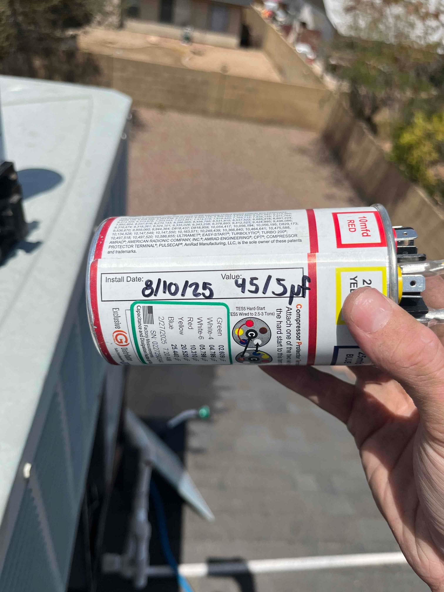 New dual-run capacitor held near the outdoor condenser, labeled with install date 8/10/25 and rated 45/5 μF.