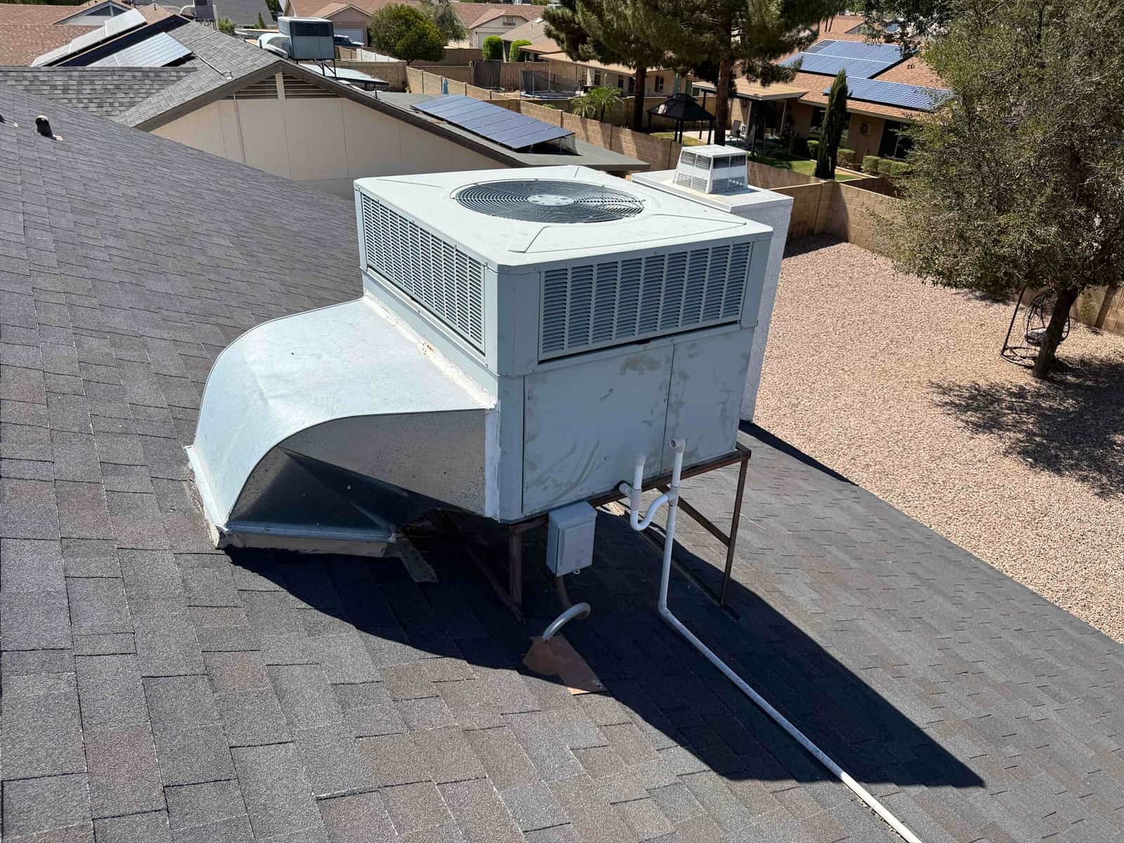 Rooftop packaged air conditioning unit on a shingle roof with sheet-metal supply duct, electrical conduit, and condensate drain piping.