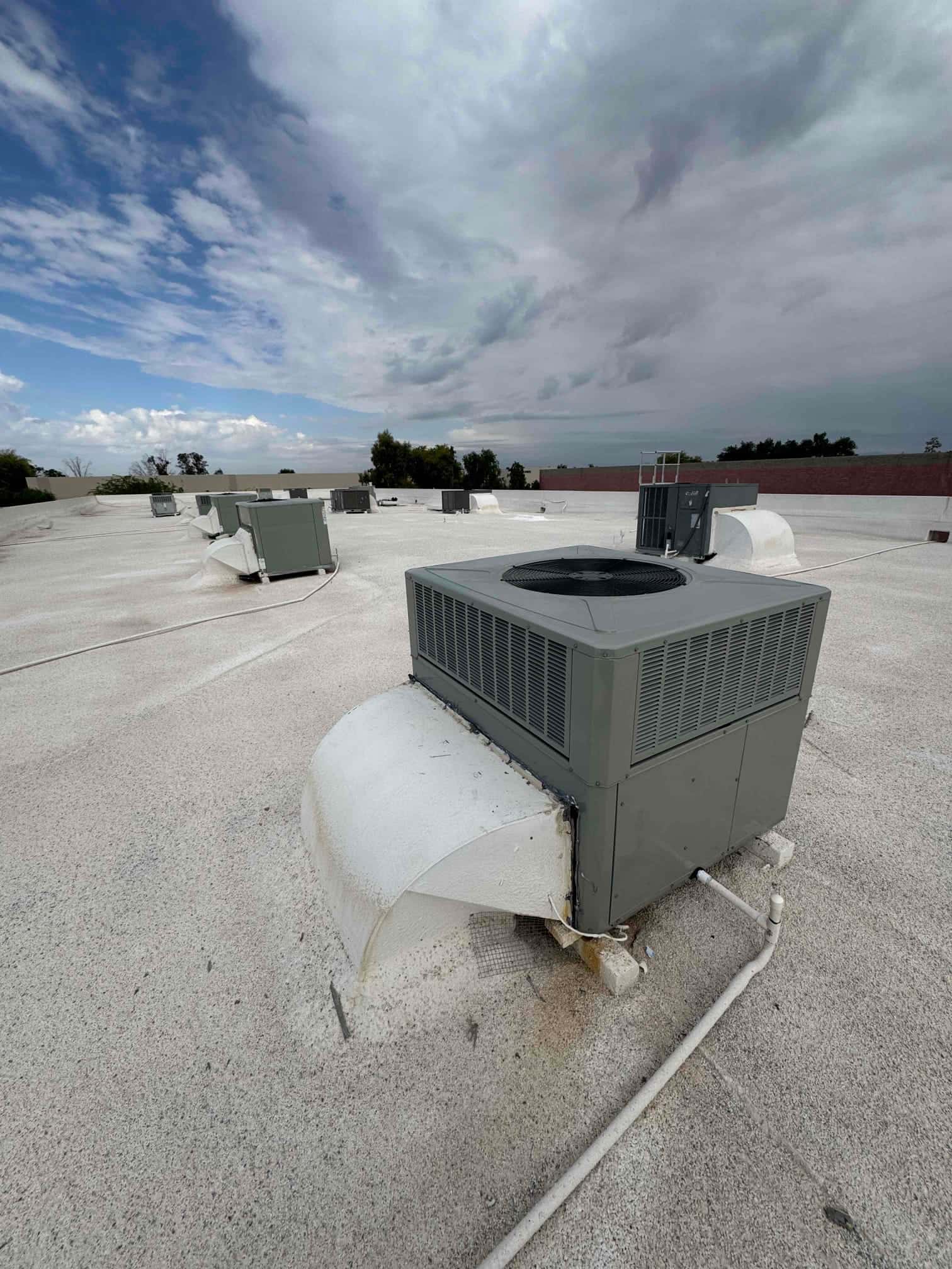 Rooftop packaged HVAC units set on curbs with connected condensate piping across a flat commercial roof under cloudy skies.