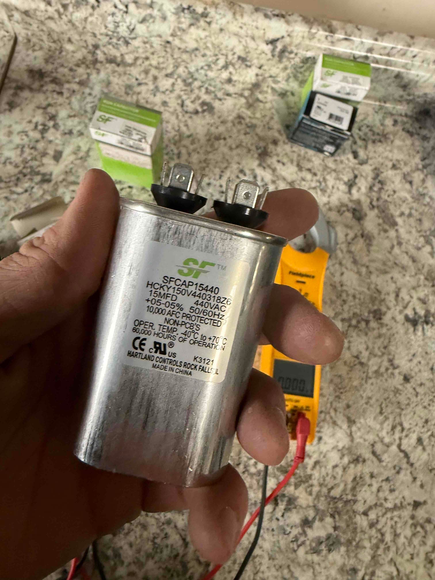 Technician holds a dual-run capacitor labeled 45/5 μF, 440/370V while a multimeter with leads sits in the background.