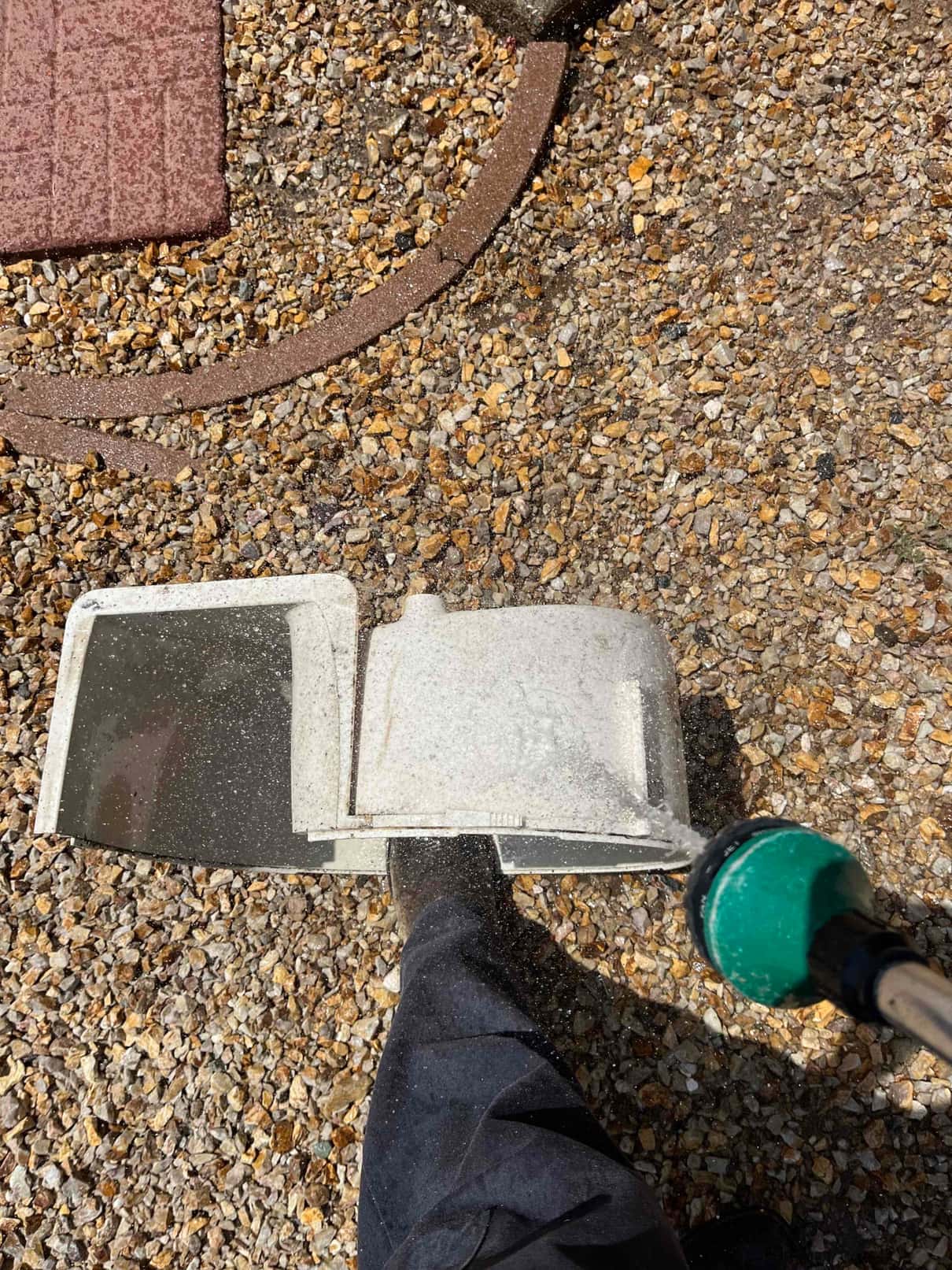 Technician rinses an HVAC air filter or panel with a hose nozzle on a gravel surface during cleaning.