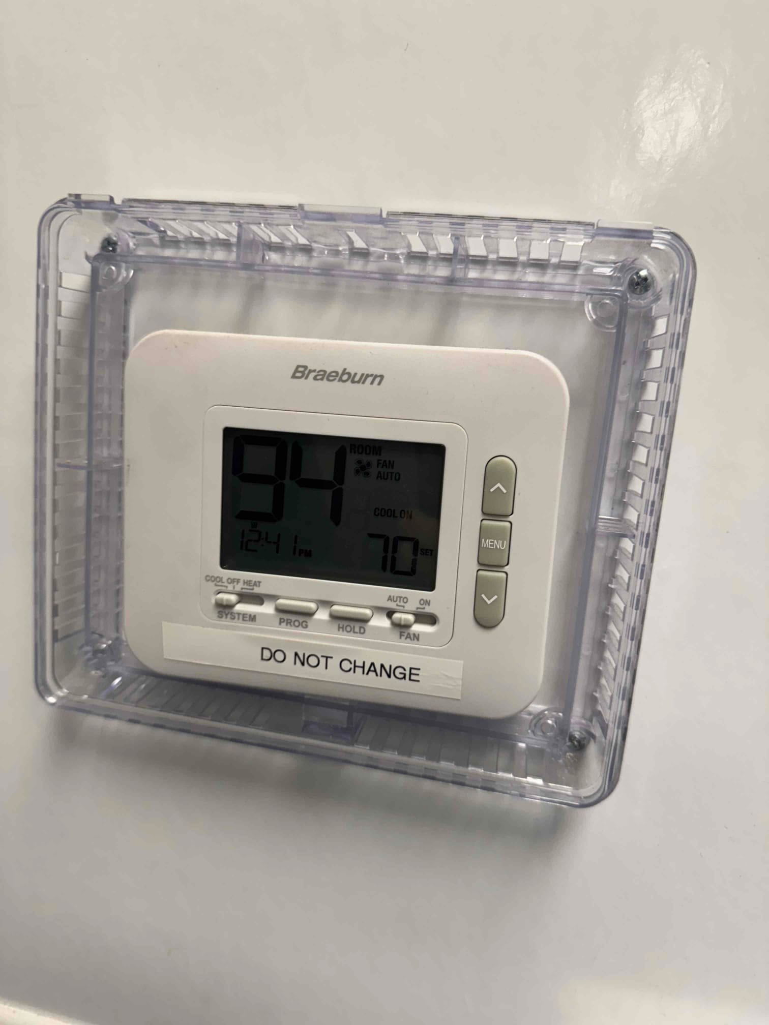 Wall-mounted digital thermostat inside a clear lockable cover, display showing indoor temperature 94 and cooling setpoint 70.