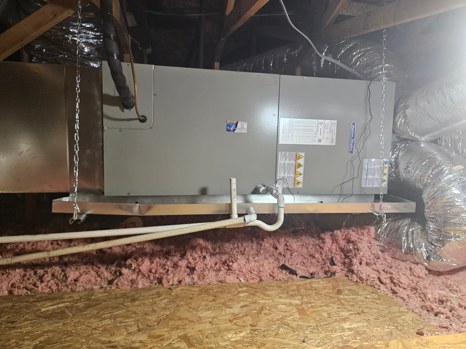 Air handler suspended in attic with insulated ducts connected, auxiliary drain pan, and PVC condensate drain piping installed.