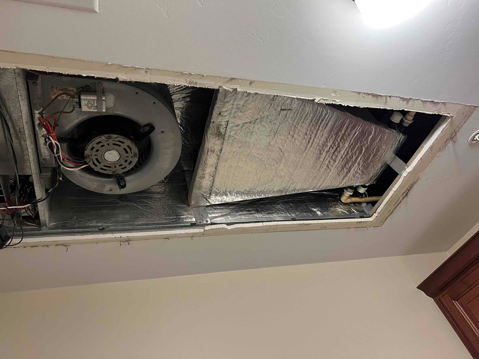 Ceiling-mounted air handler with blower motor and insulated evaporator cabinet exposed during installation, showing wiring and condensate drain connections.