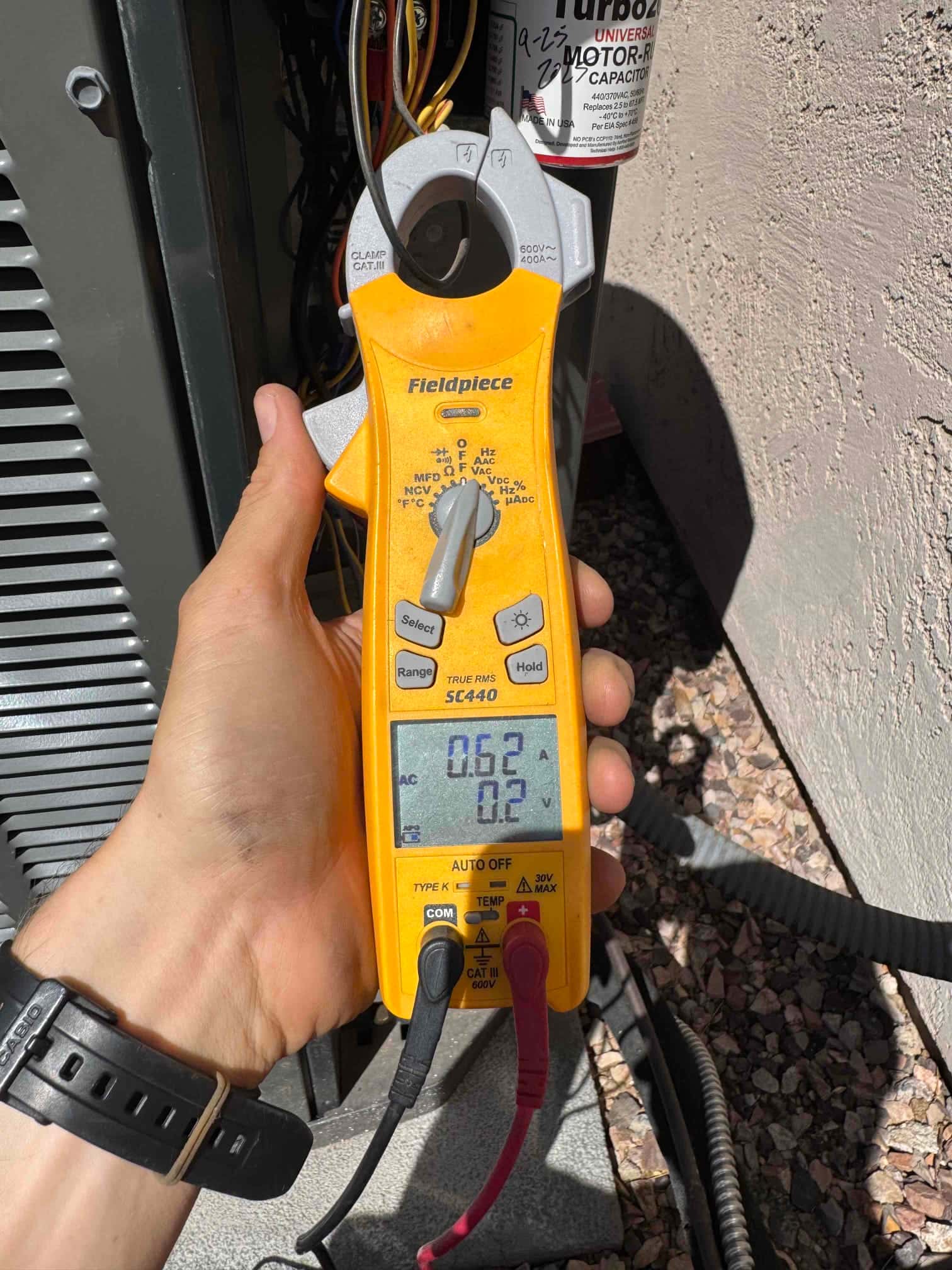 Clamp meter measures 0.62 A on a conductor at the outdoor condenser, with test leads connected and display clearly visible.