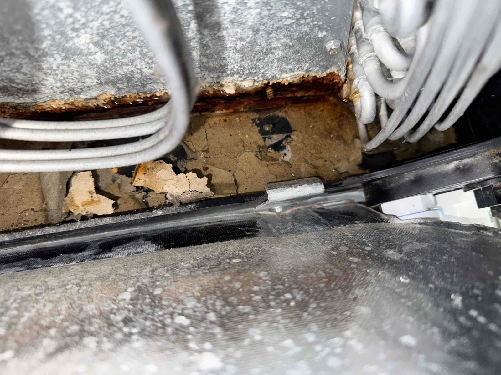 Condensate drain pan area shows rust and flaking debris beneath evaporator coil tubing, indicating corrosion and accumulated sediment around the drain path.