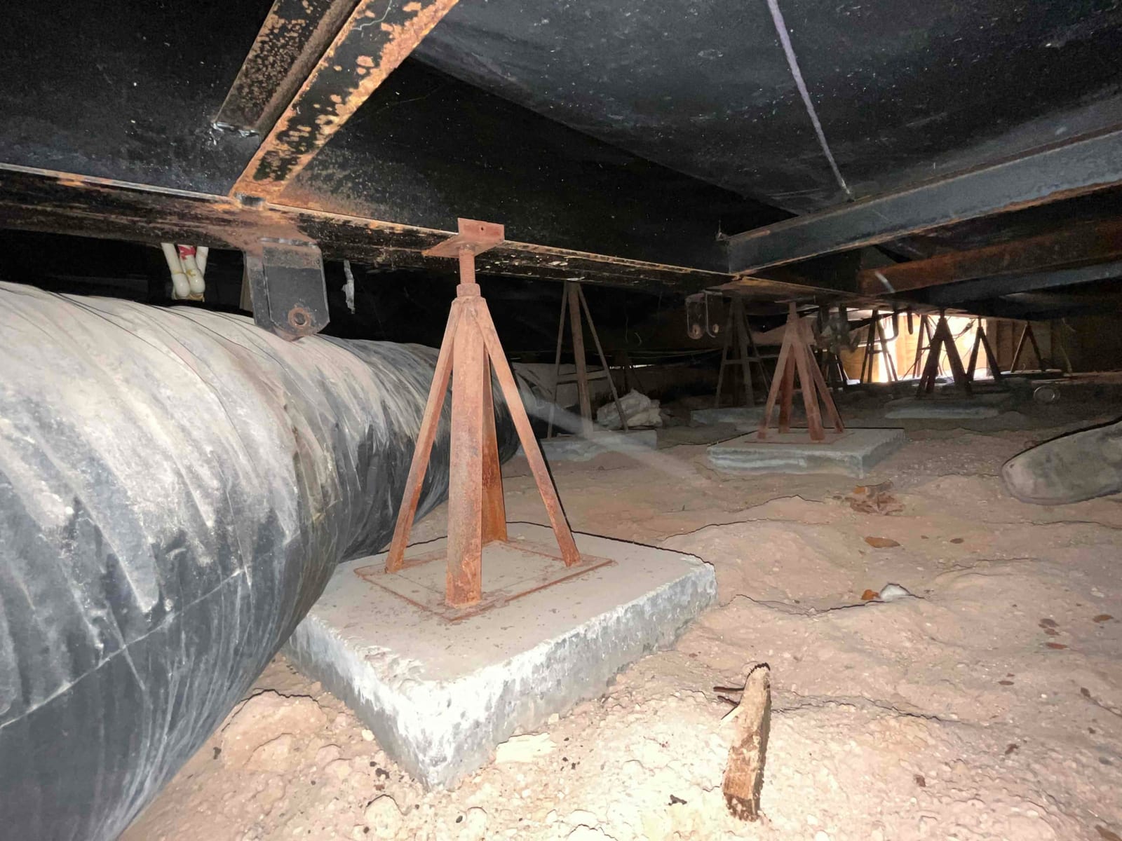 Crawlspace view showing large flexible duct and multiple rusted support stands on concrete pads.
