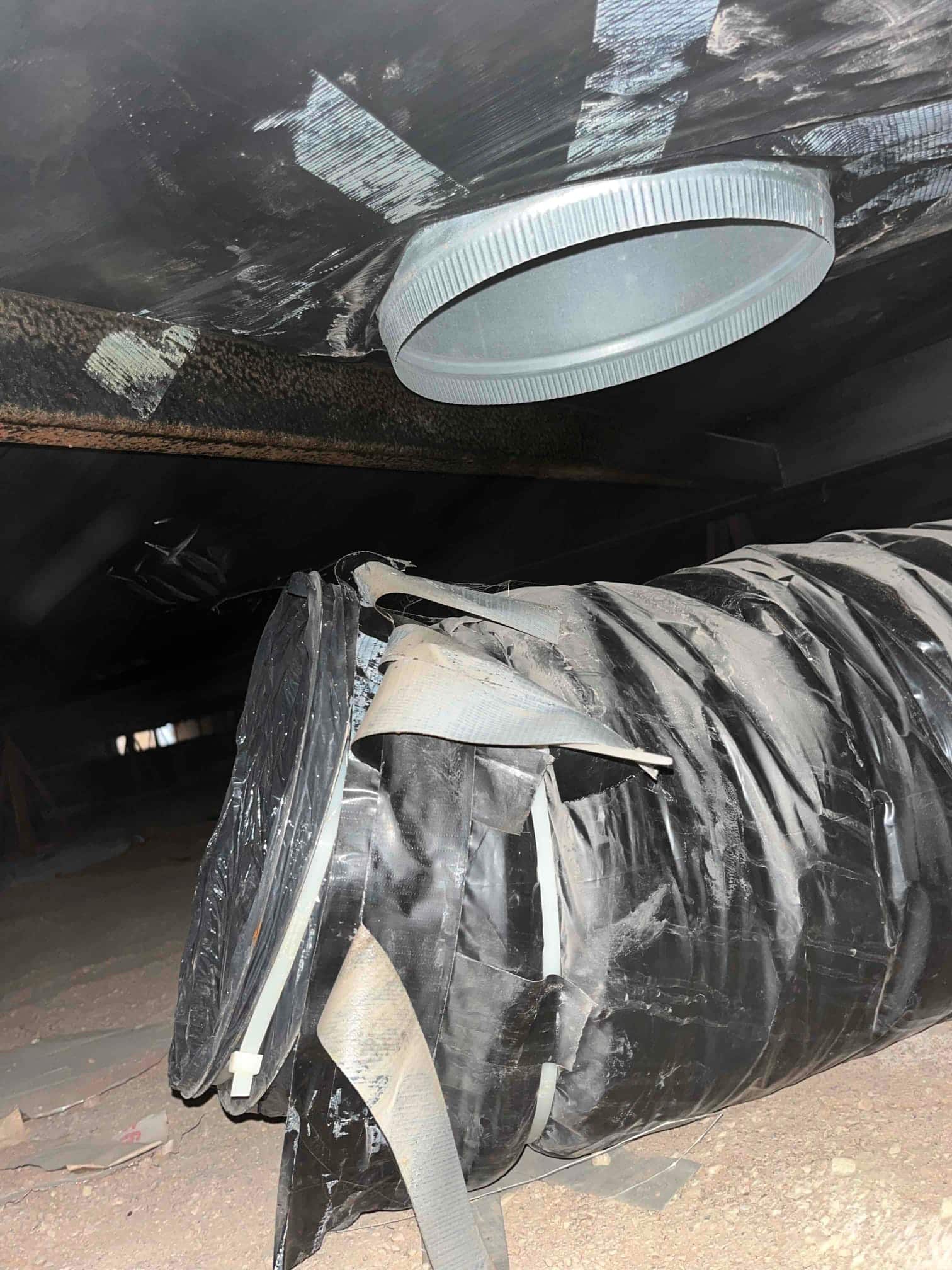 Damaged flexible duct with torn outer jacket lies on the floor beneath a metal takeoff in an attic space.
