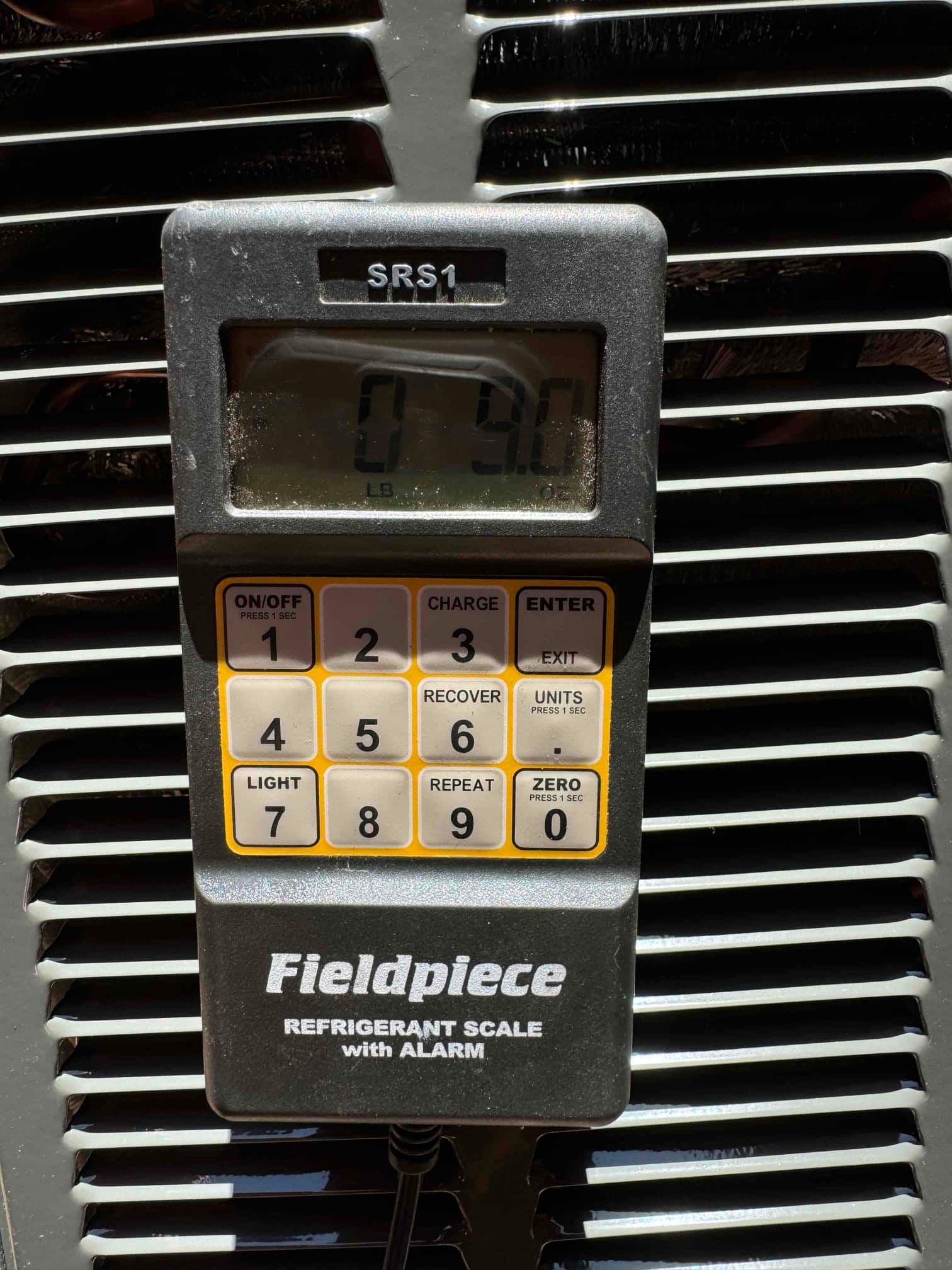 Digital refrigerant scale controller resting on the outdoor condenser grille, display showing weight in pounds and ounces.