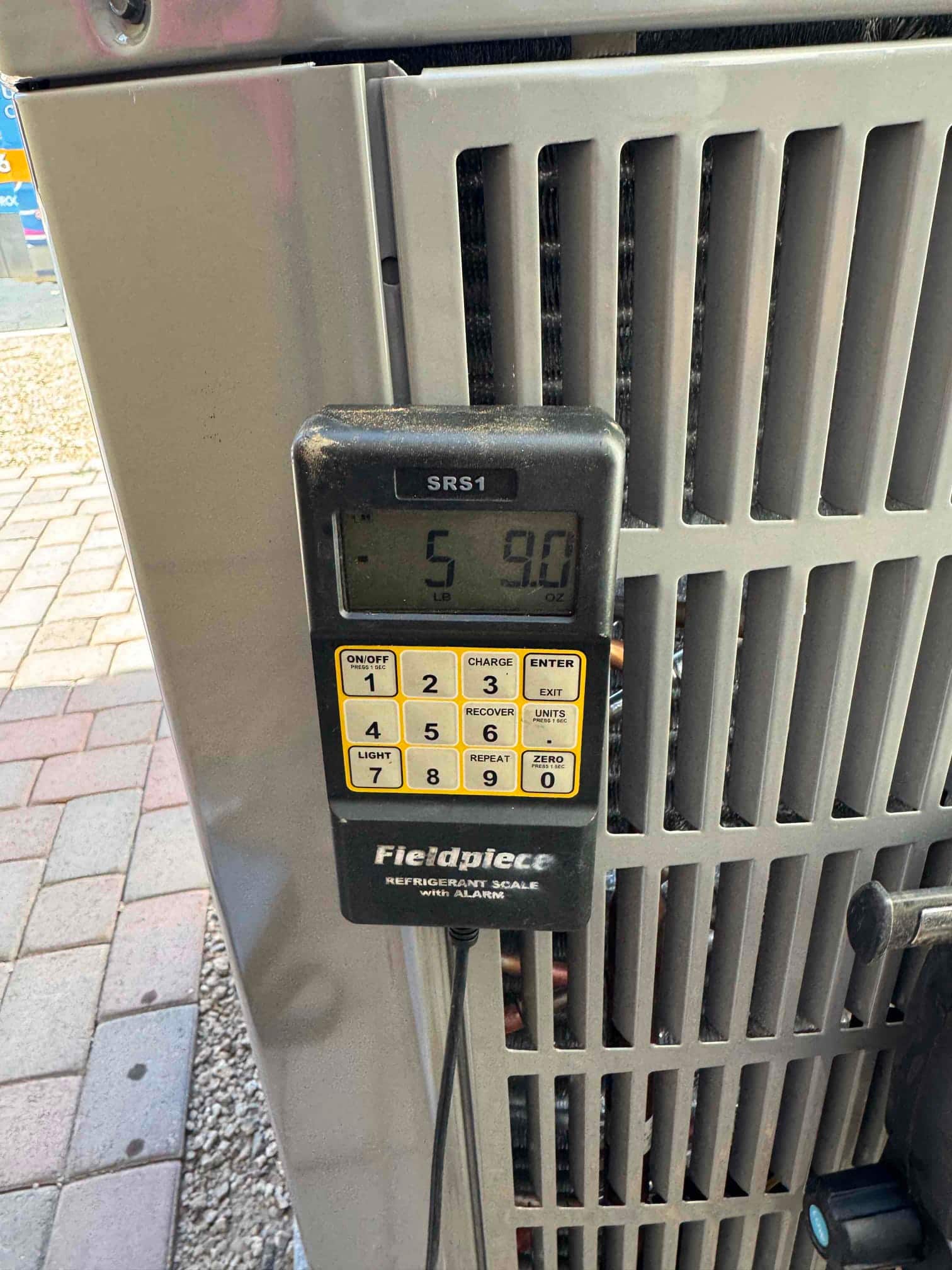 Digital refrigerant scale displays 5 pounds 9 ounces during charging beside the outdoor condenser cabinet.