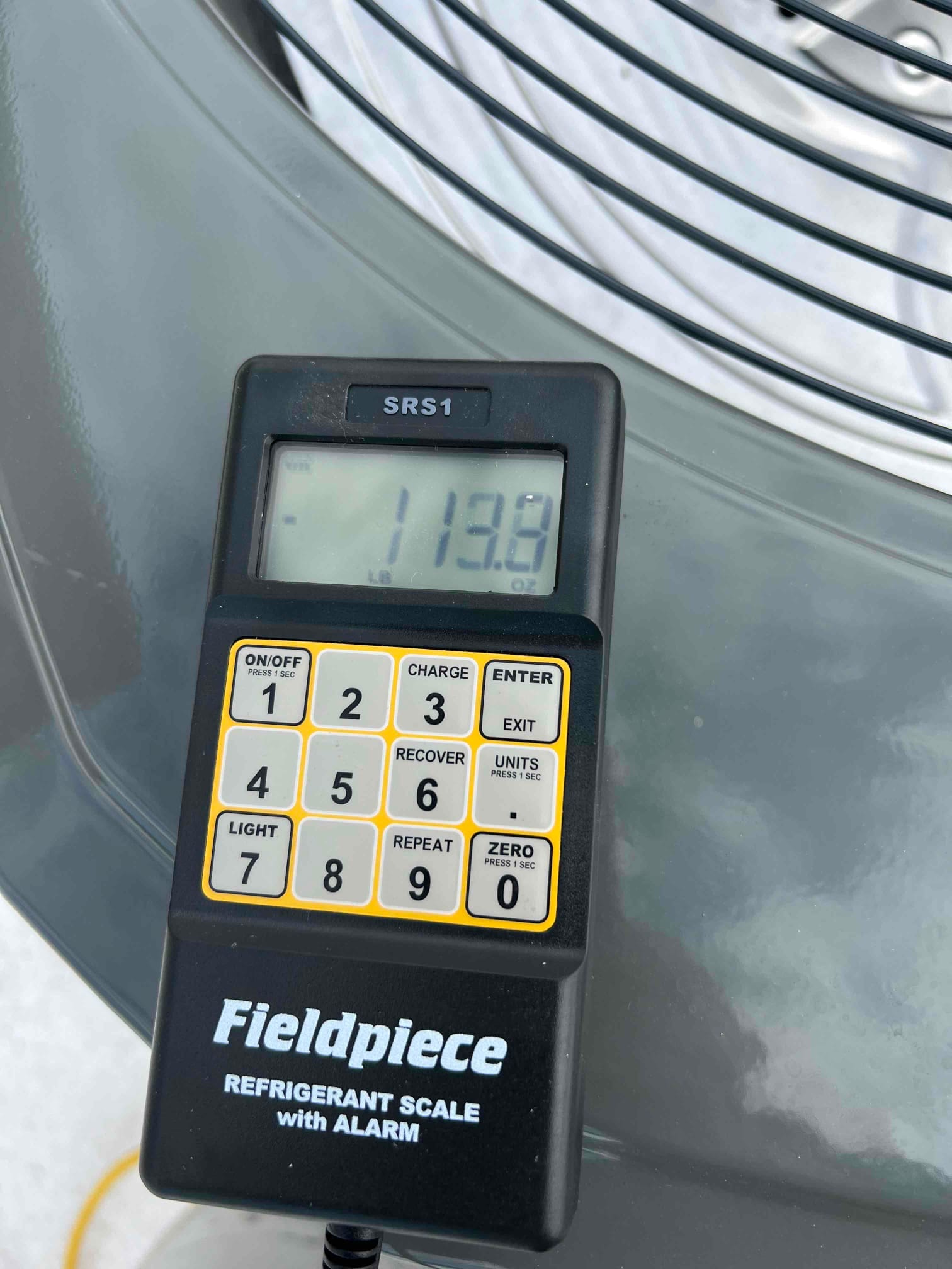 Digital refrigerant scale handset displays 113.8, showing charged weight during installation beside the outdoor condenser fan guard.