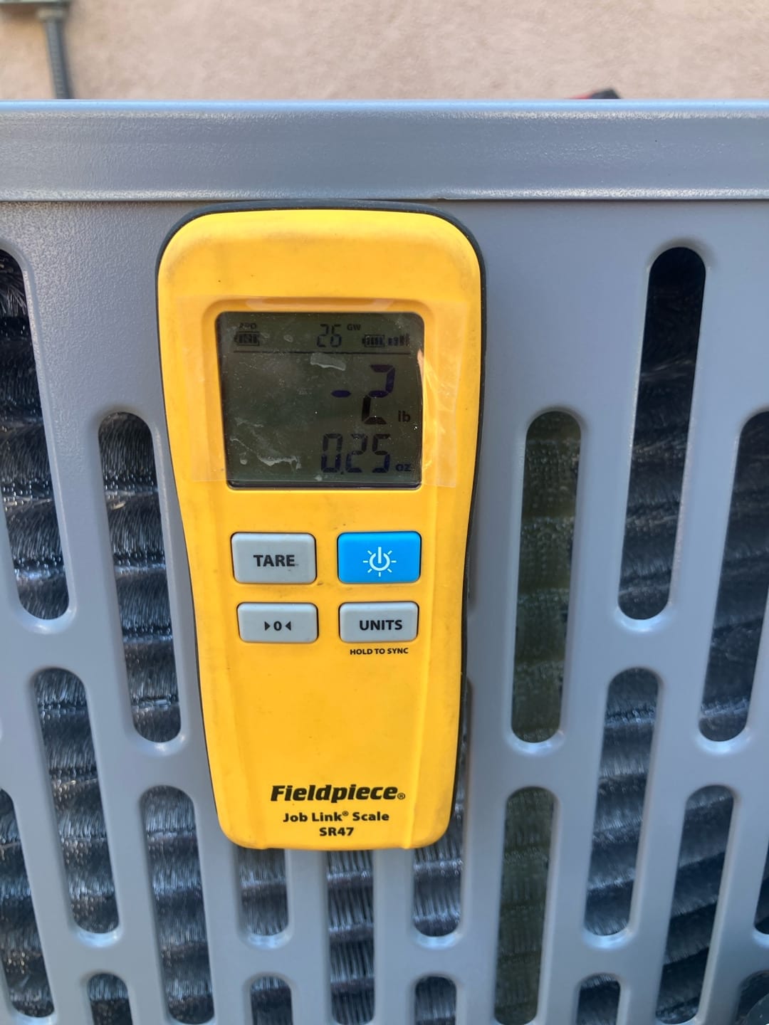 Digital refrigerant scale hanging on the outdoor condenser grille, displaying a weight reading during refrigerant handling.