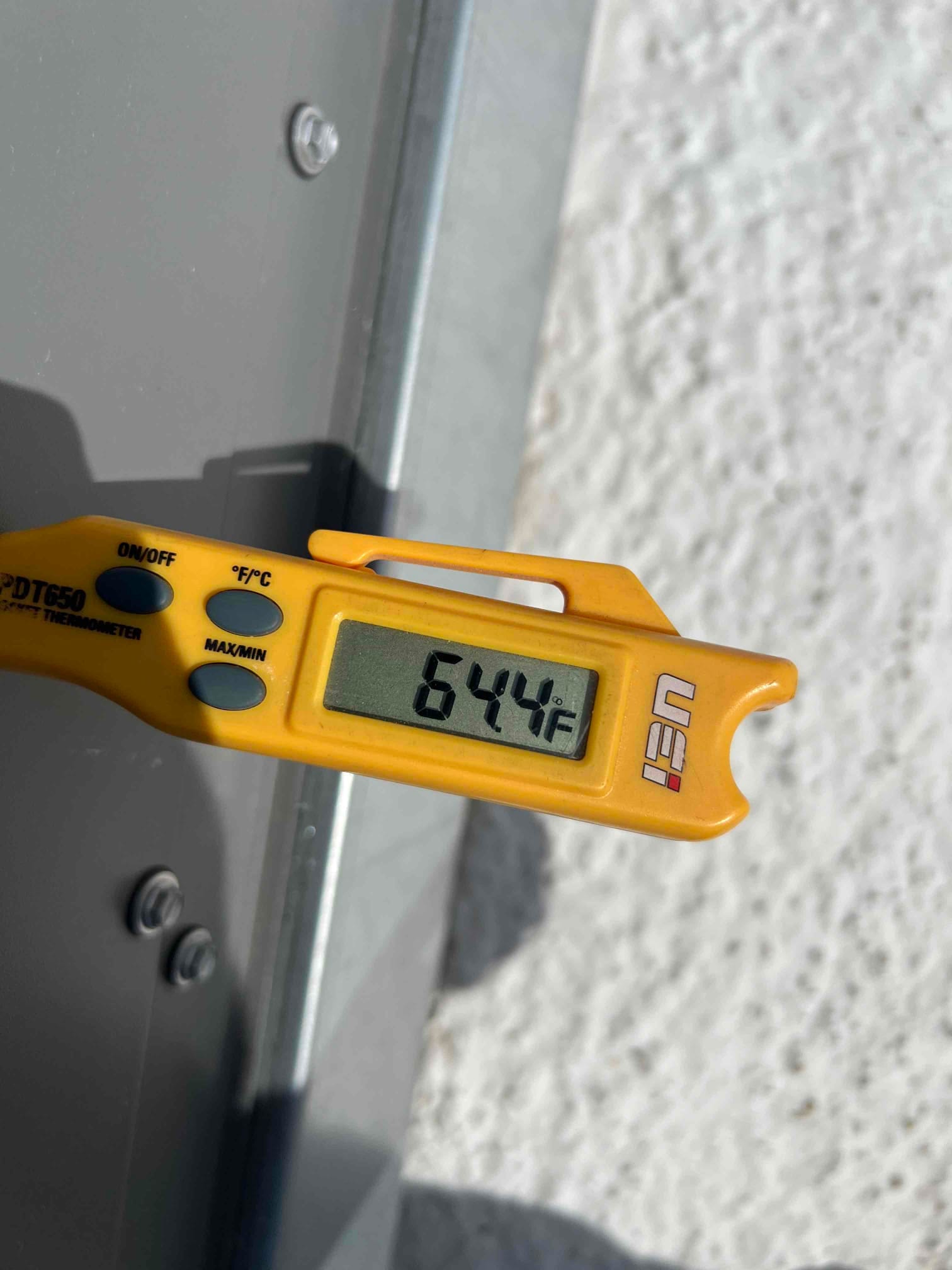 Digital thermometer displays 64.4°F while checking air temperature during commissioning at the outdoor unit.