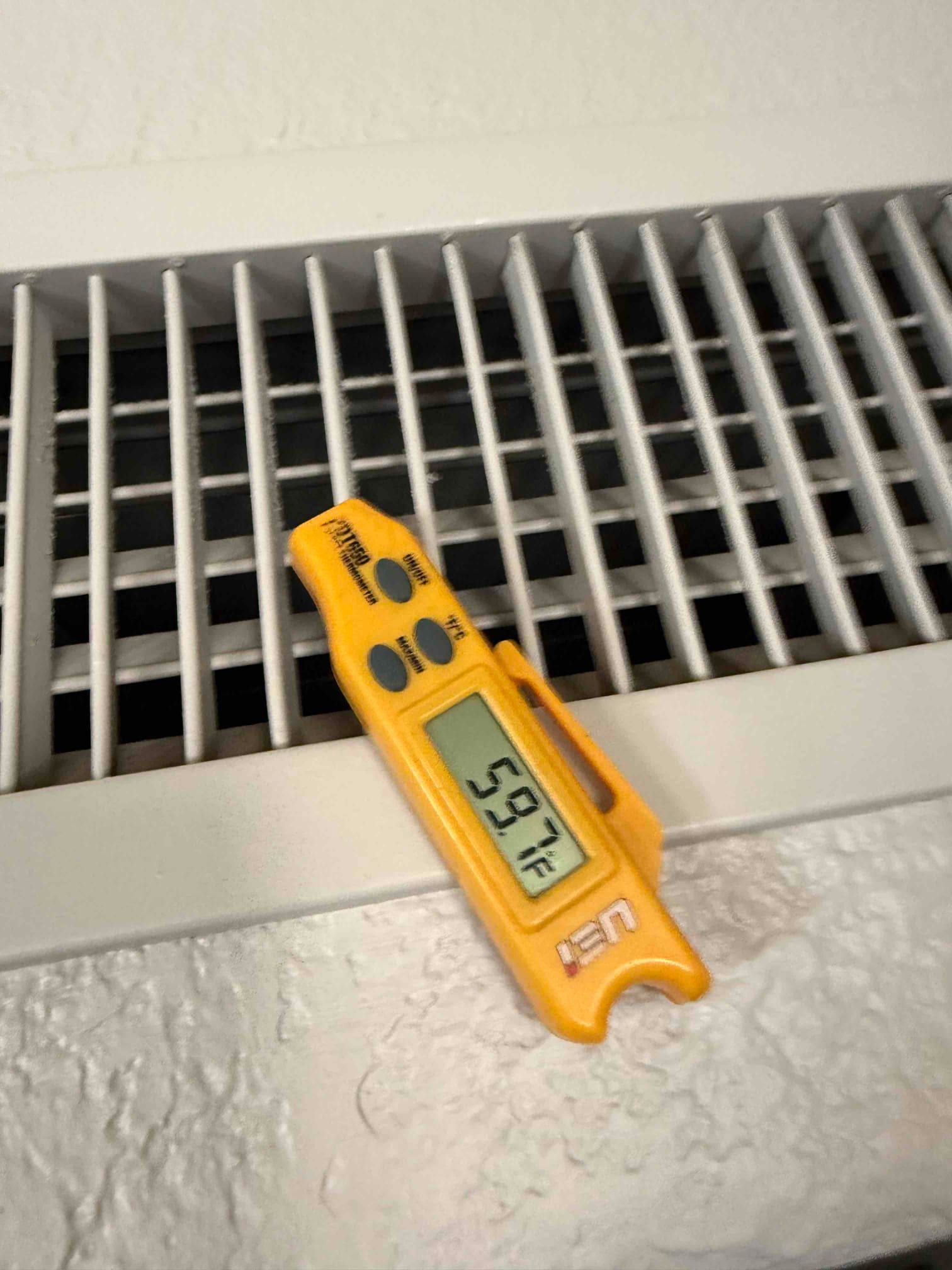 Digital thermometer rests on a supply vent grille, showing 59.7 °F supply air temperature from the register.