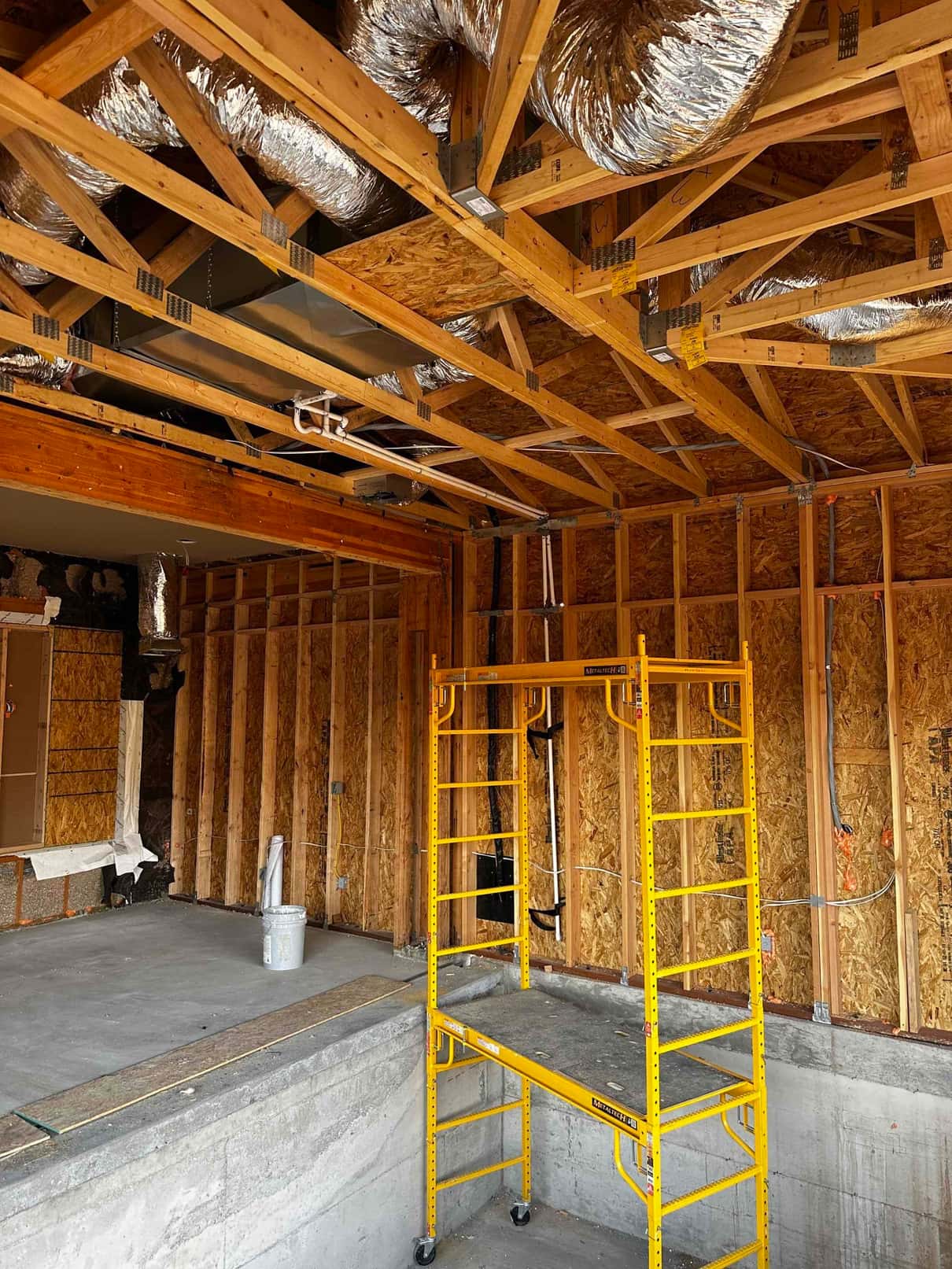 Ductwork and an air handler plenum installed within open framing, with insulated flexible ducts routed above and a work scaffold below.