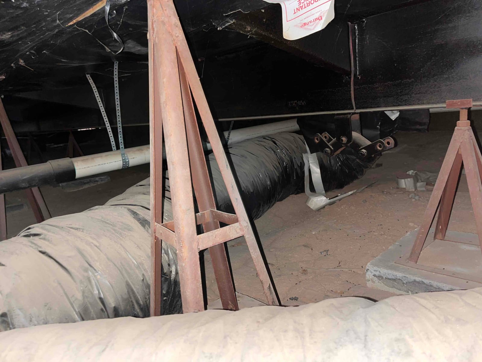 Flexible insulated duct run in a crawlspace supported by metal stands, with adjacent refrigerant and condensate lines visible.