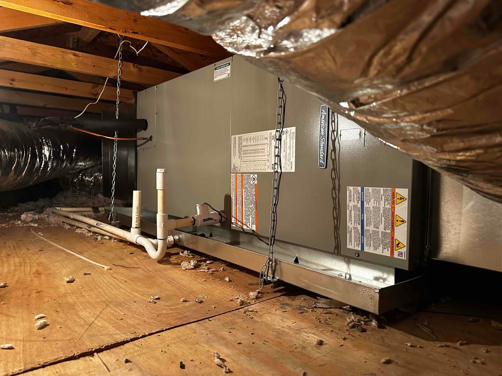 Horizontal air handler installed in attic with suspended support chains, insulated duct connections, and PVC condensate drain routed to a safety pan.