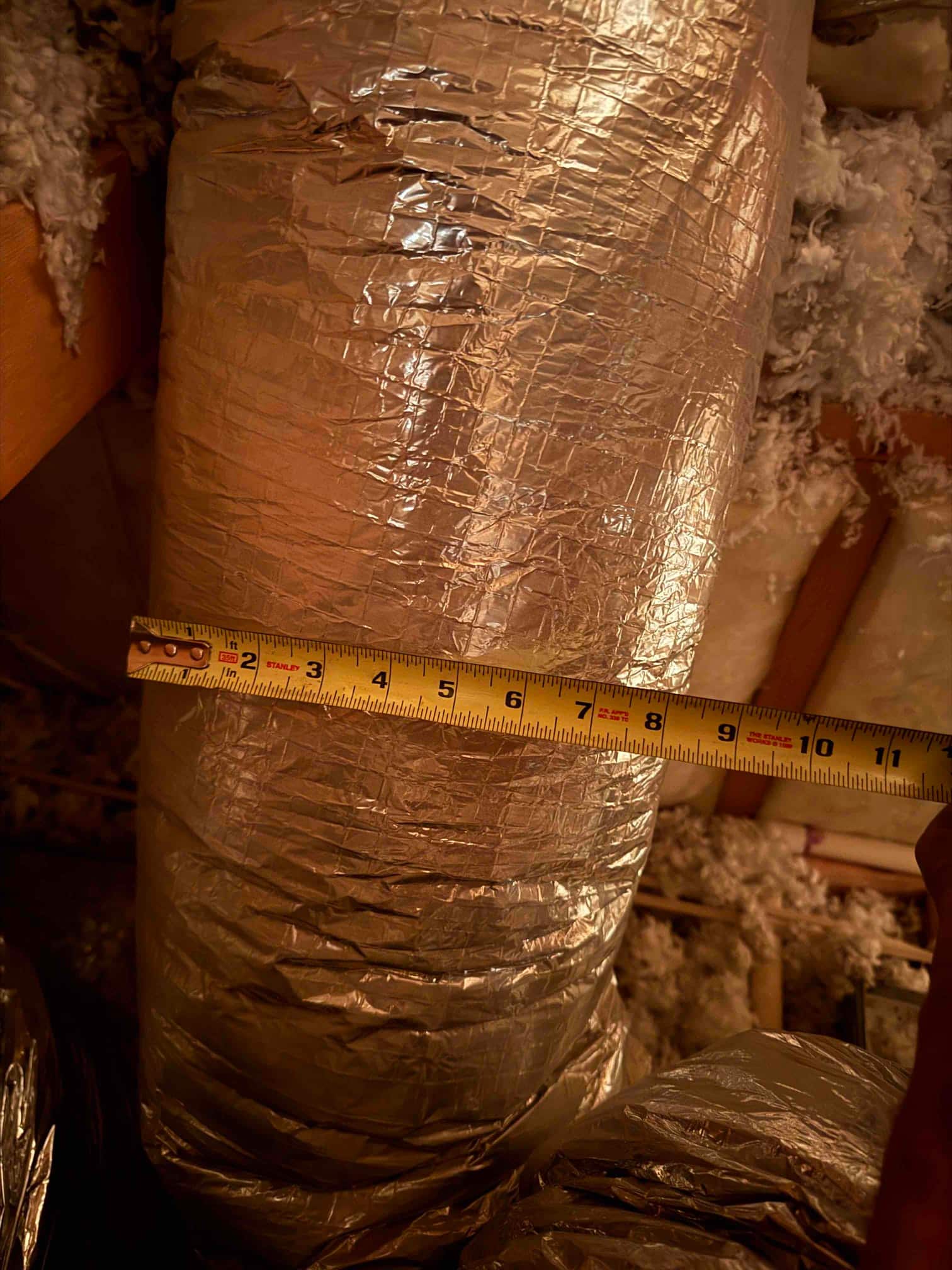 Measuring the diameter of insulated flexible duct in an attic using a tape measure for sizing verification.