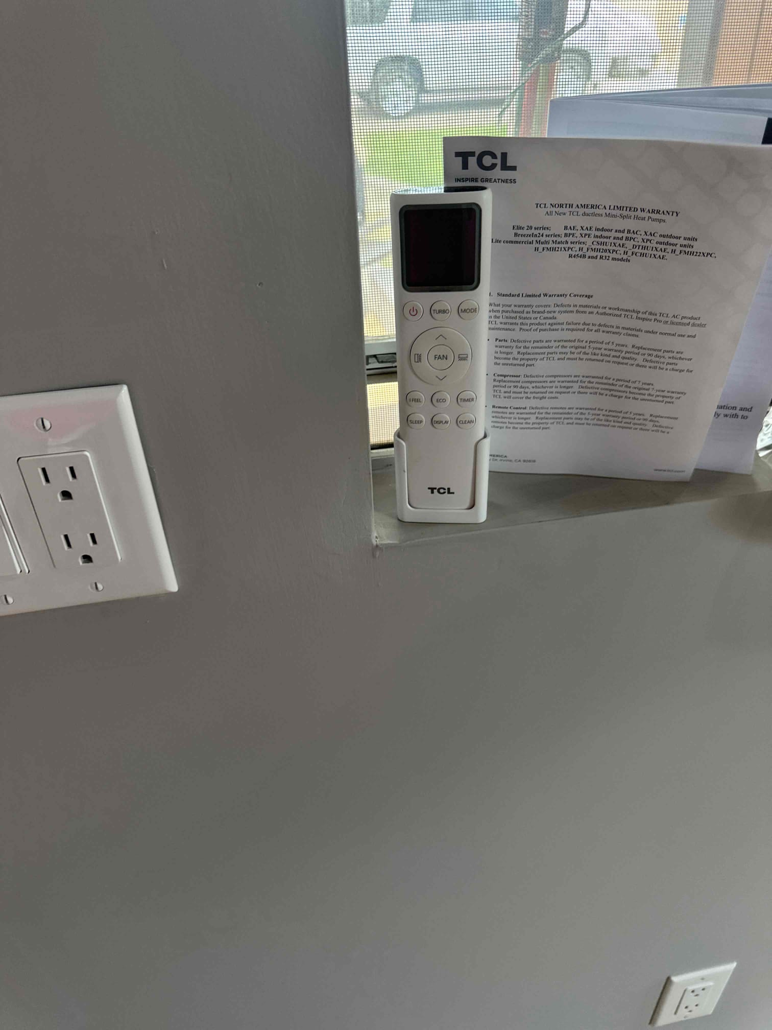 Mini-split handheld remote placed on an interior windowsill beside a wall outlet and warranty document after installation.
