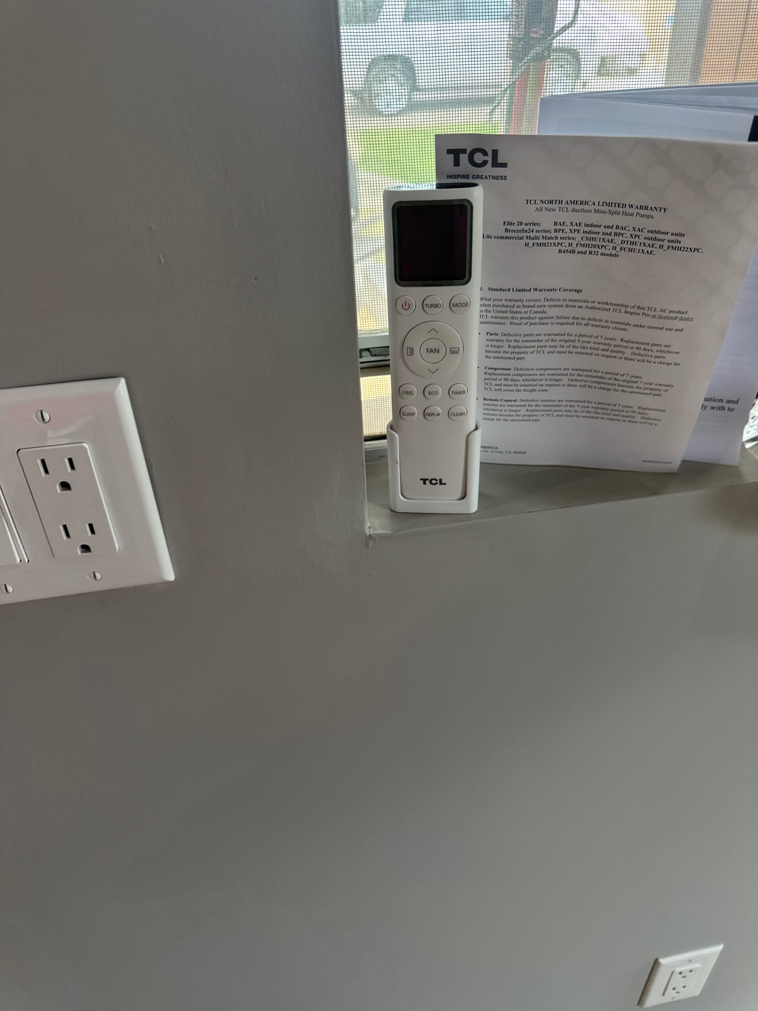 Mini-split handheld remote placed by the window after installation, with wall outlets and warranty paperwork visible nearby.