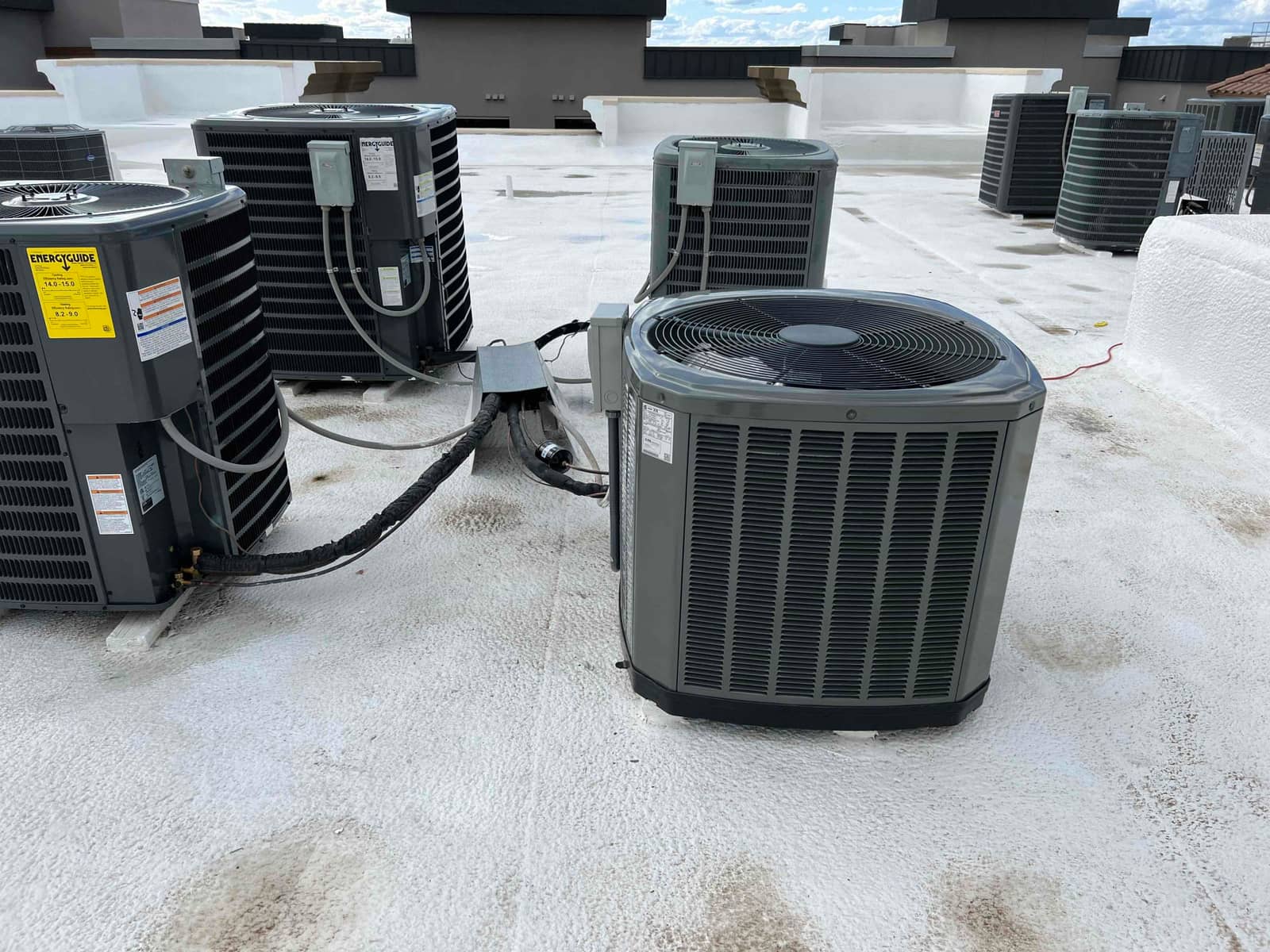Multiple rooftop condensers installed on a coated roof with insulated refrigerant lines and electrical disconnects visible beside the units.