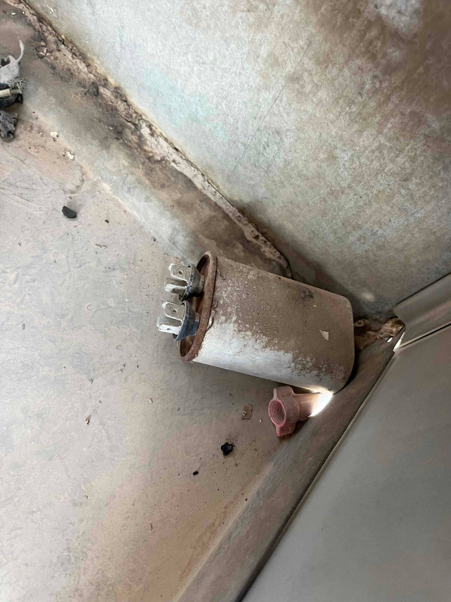 Old dual-run capacitor removed from the condenser compartment, showing corroded case and exposed spade terminals on the top.