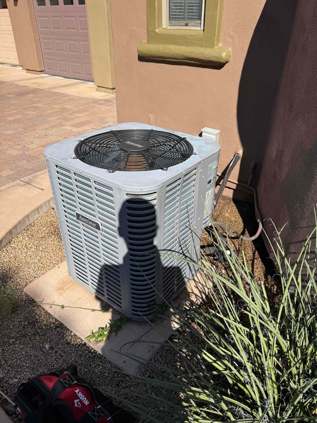 Outdoor air conditioning condenser unit beside a house, with refrigerant and electrical lines entering the wall near the base.