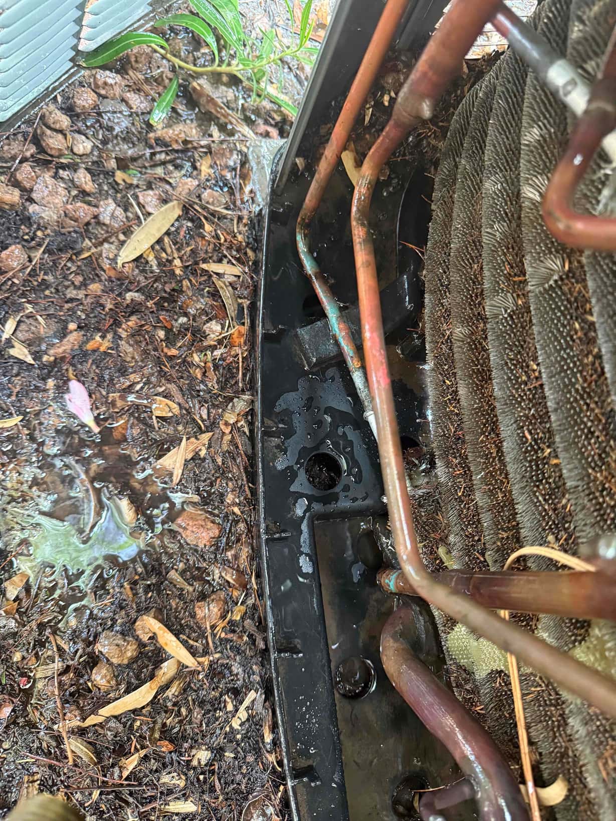 Outdoor condenser coil and copper refrigerant lines exposed, with wet debris and water pooling in the base pan near the tubing.