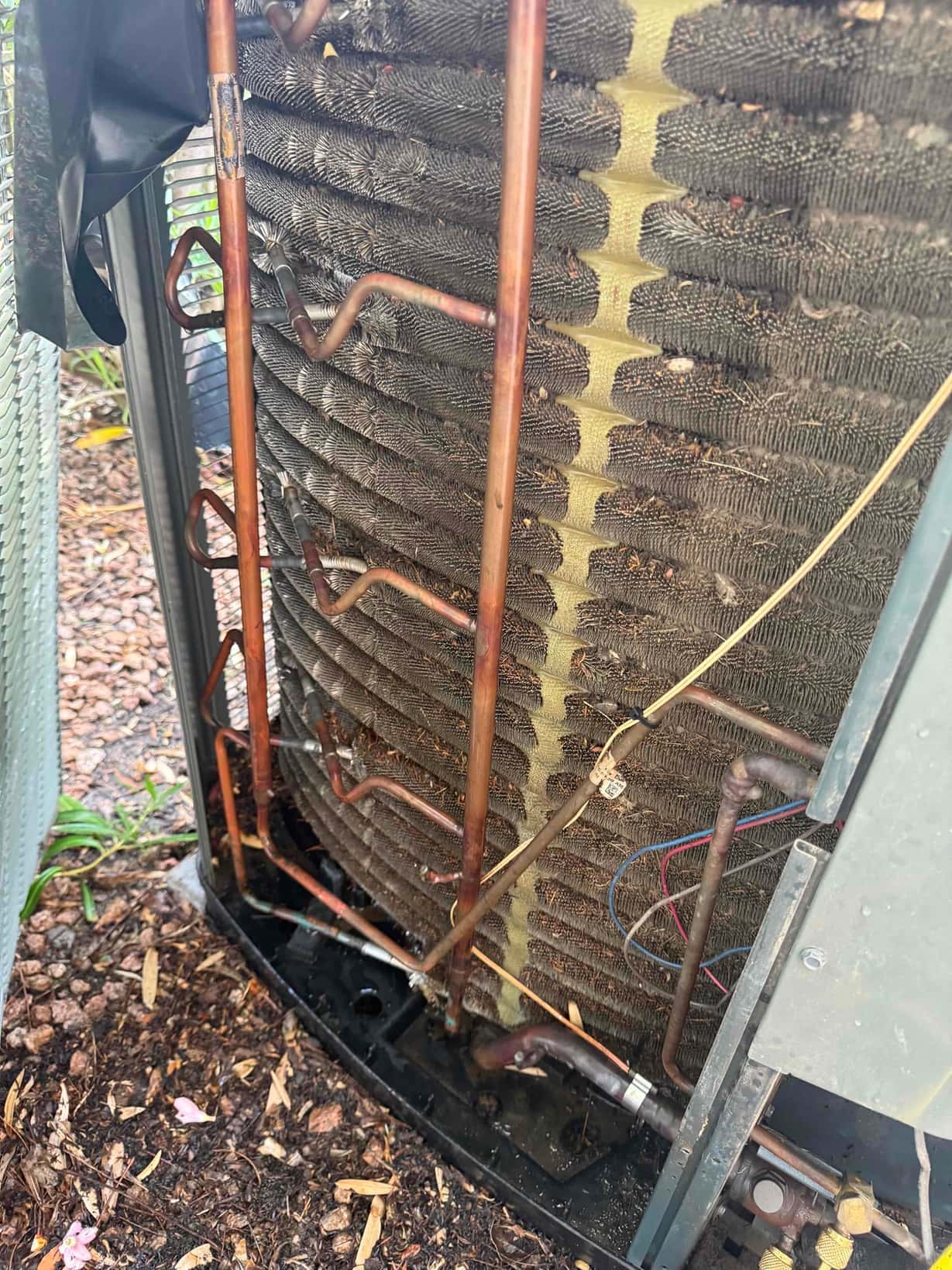 Outdoor condenser coil with exposed copper refrigerant tubing and wiring, showing dirt and debris at the unit base.