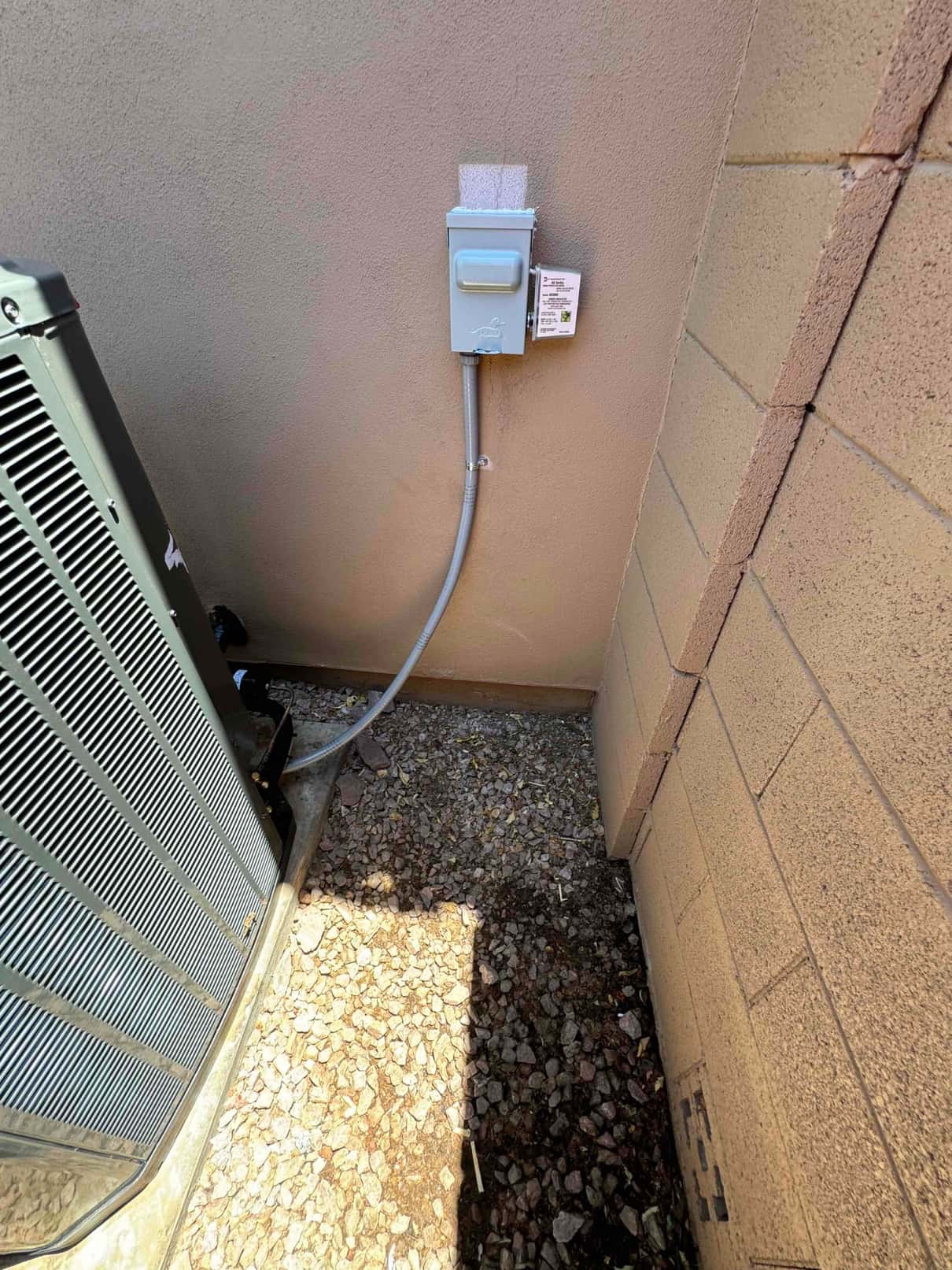 Outdoor condenser next to a wall with newly installed weatherproof disconnect and flexible conduit routed to the unit.