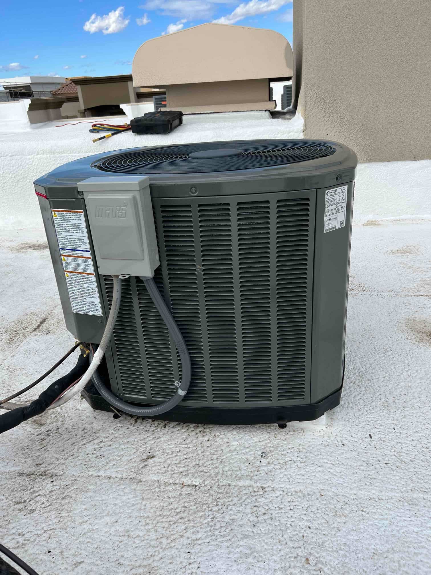Outdoor condenser set on a rooftop surface with connected refrigerant lines, electrical whip, and nearby service disconnect.