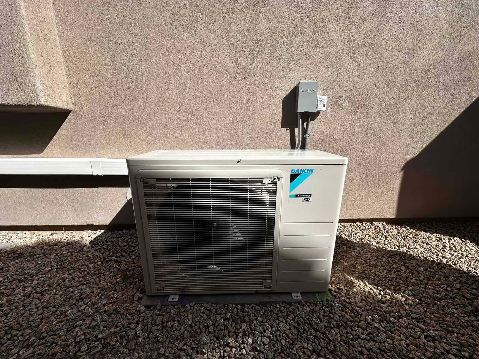 Outdoor mini-split condenser set on a level pad with line set cover and mounted electrical disconnect on the wall.