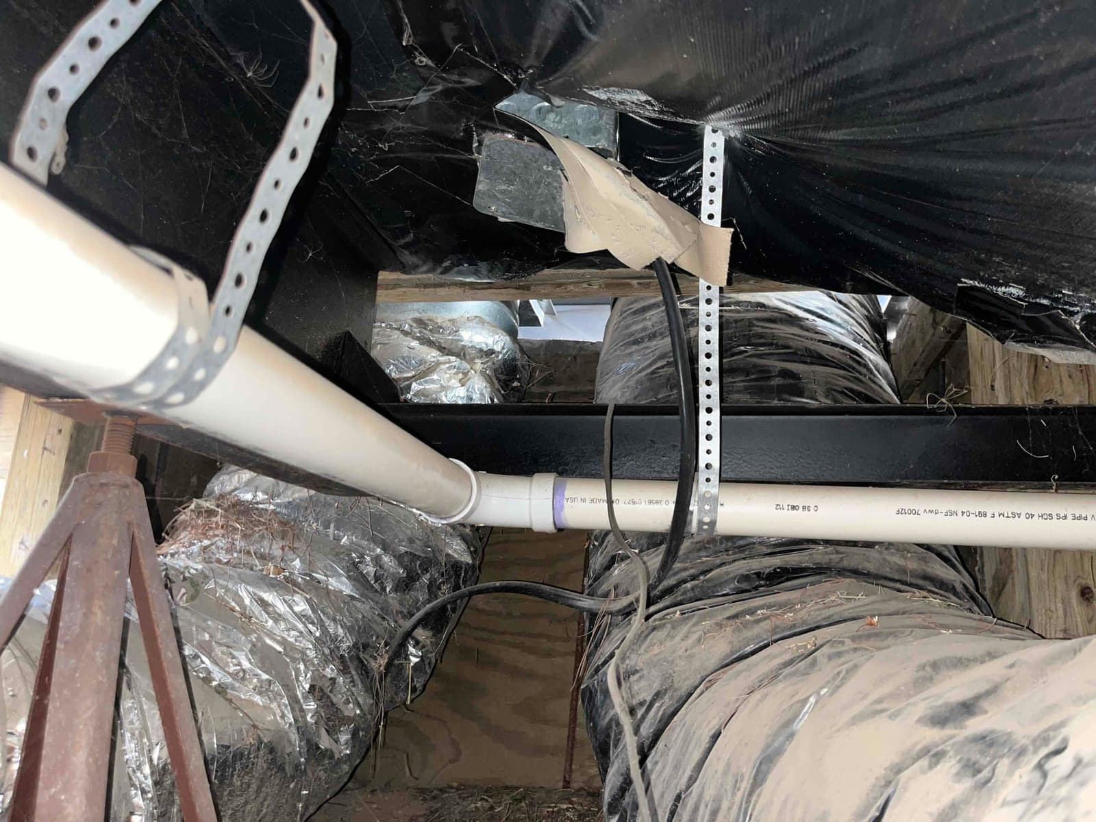 PVC condensate drain runs through attic beside insulated flex duct and support straps, showing wiring and surrounding framing.