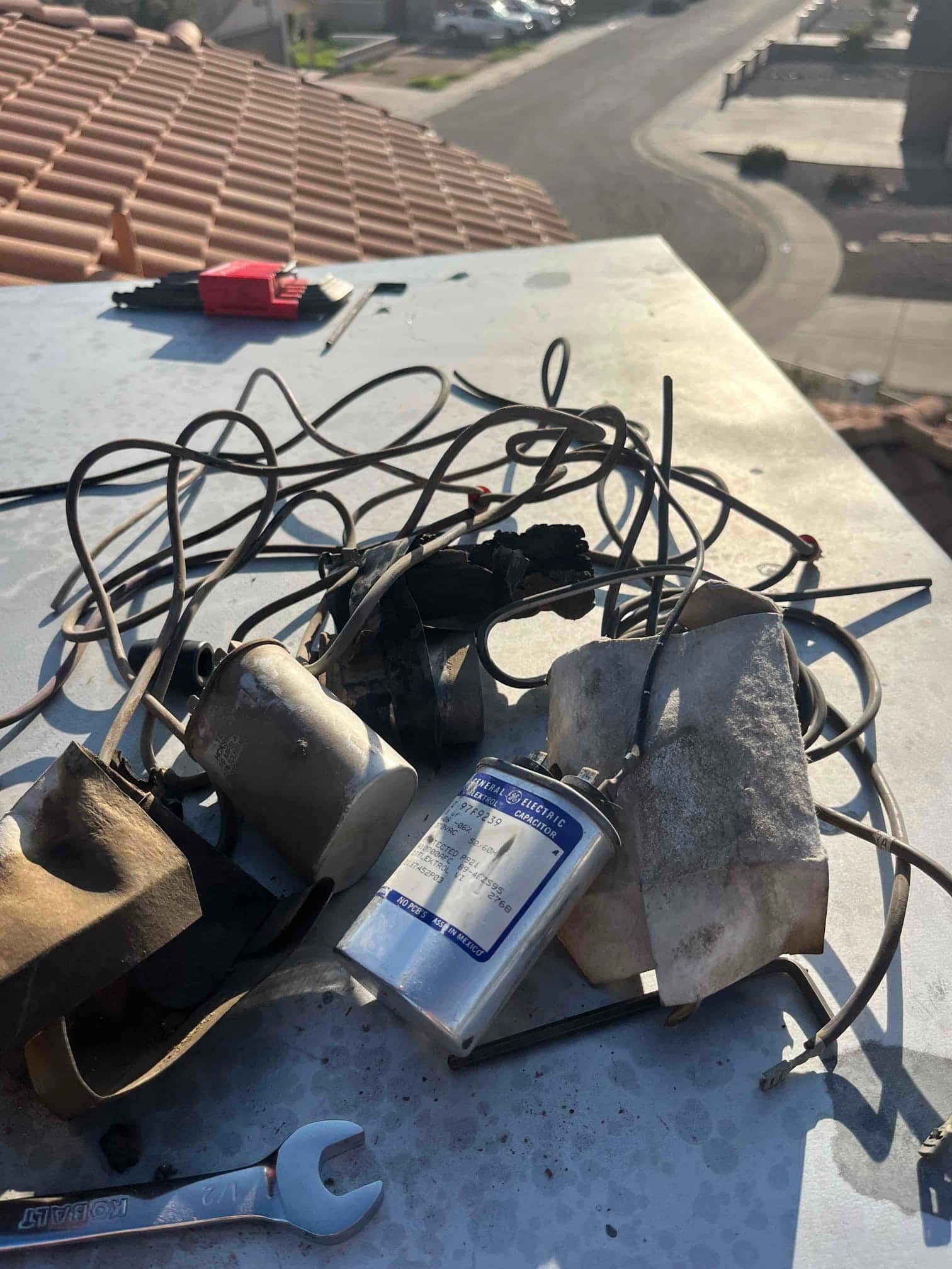Removed condenser components on a rooftop unit, including multiple run capacitors and wiring, alongside a wrench and disconnected leads.