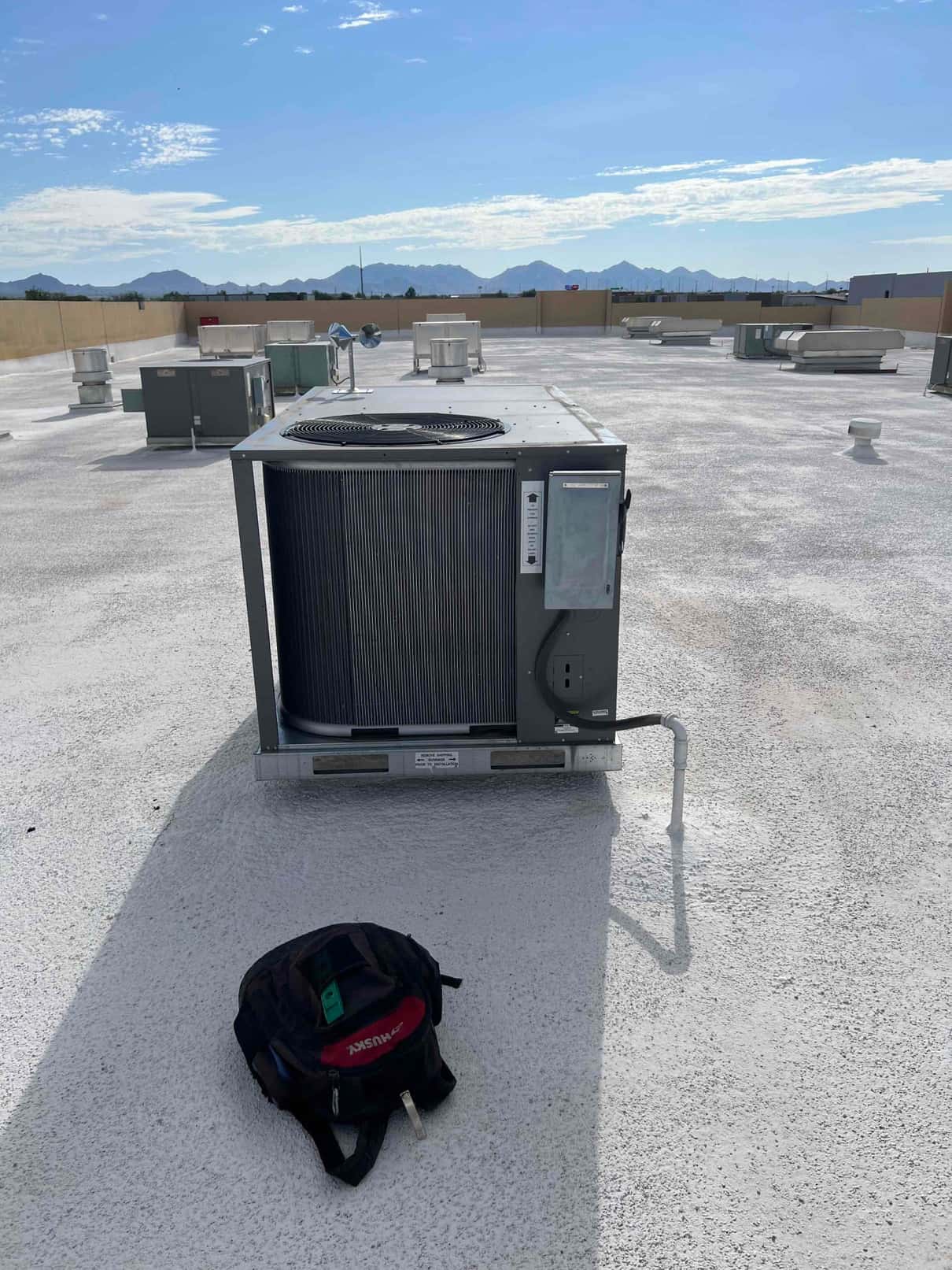 Rooftop HVAC package unit set on curb with connected condensate drain, surrounded by other rooftop equipment under clear skies.