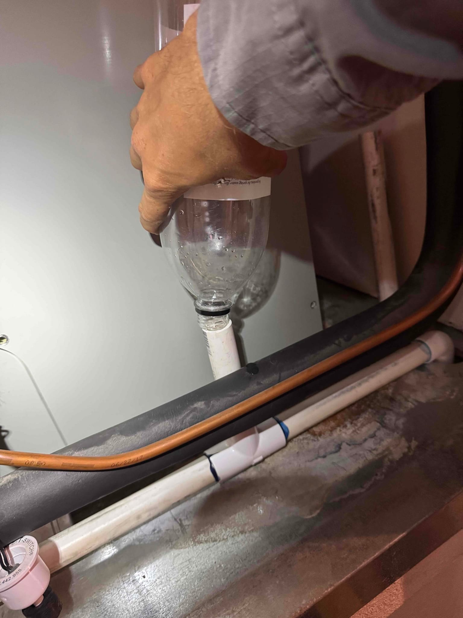 Technician flushes the condensate drain line using a plastic bottle connected to the PVC drain at the air handler.