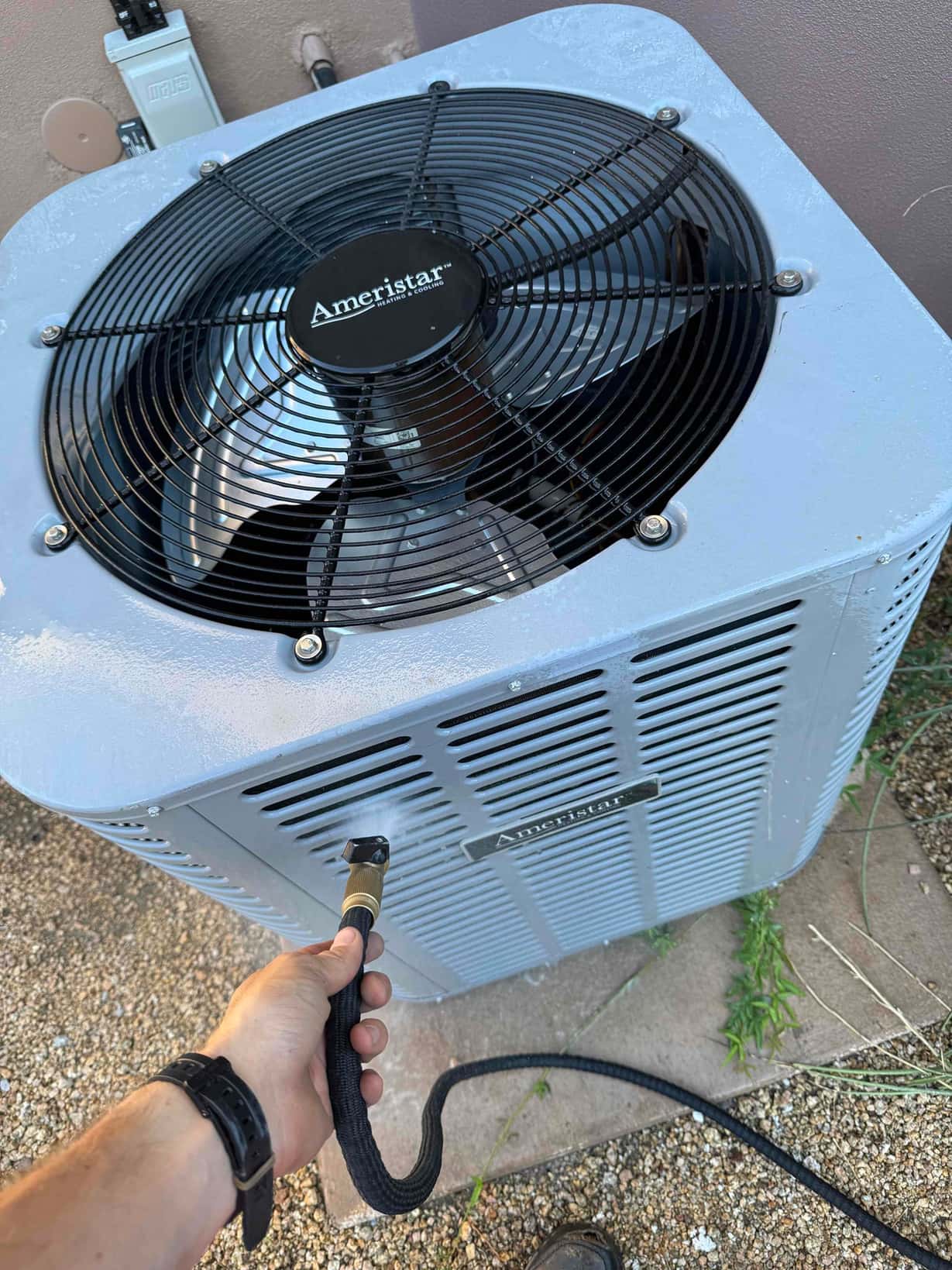 Technician flushes the outdoor condenser’s condensate drain line using a hose while the unit is installed beside a wall.