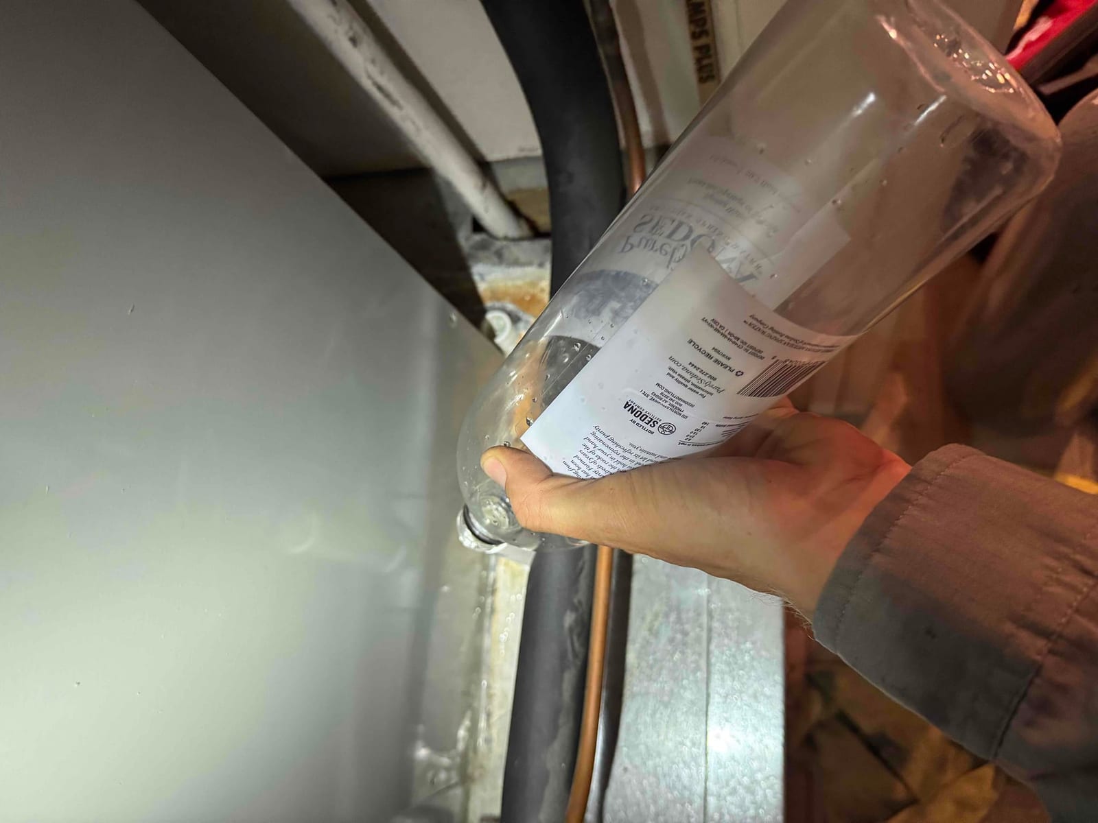 Technician’s hand flushes an HVAC condensate drain line using a clear plastic bottle at the air handler.