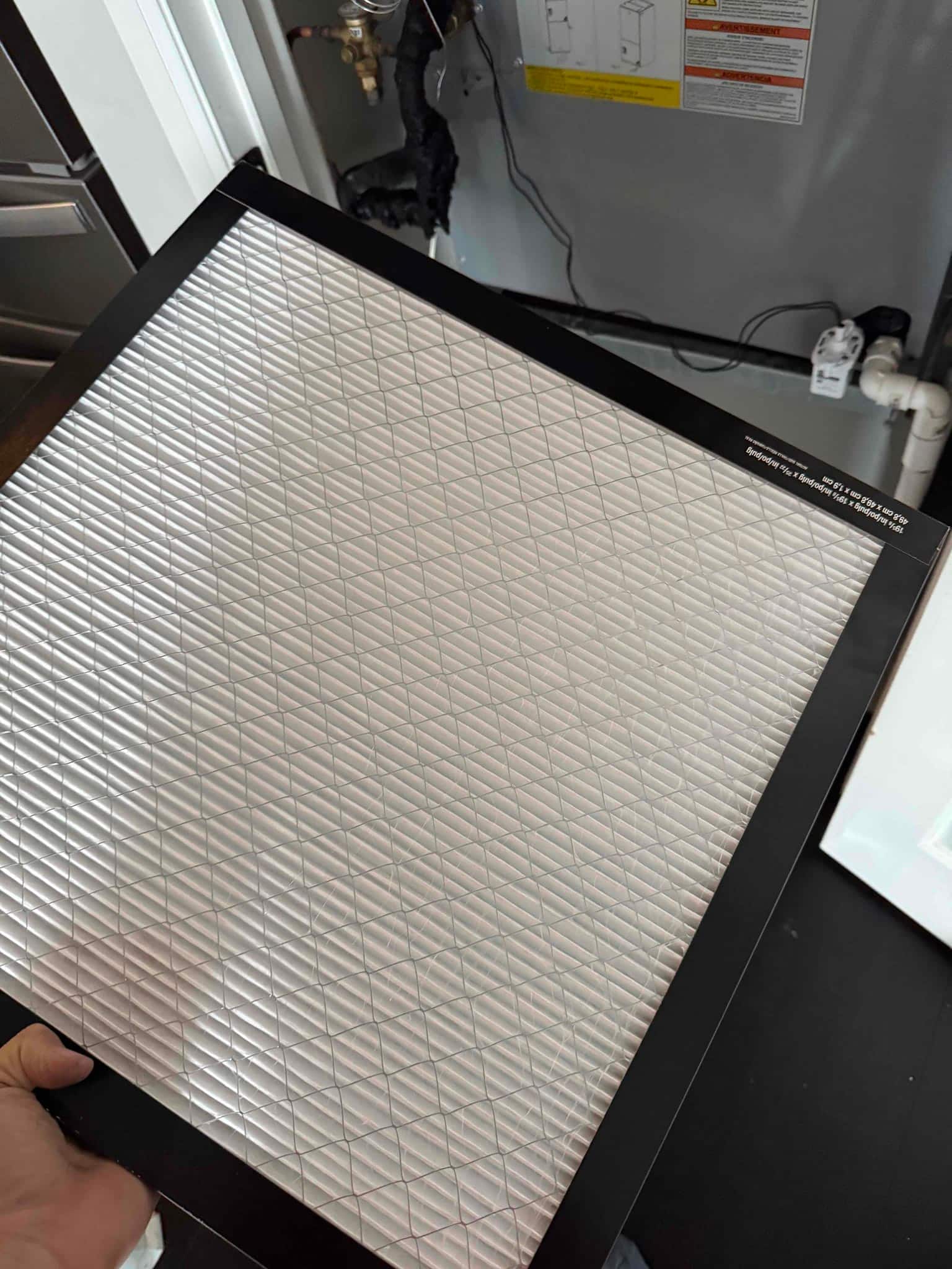 Technician holds a clean air filter near an indoor air handler with visible PVC condensate drain and service panel.